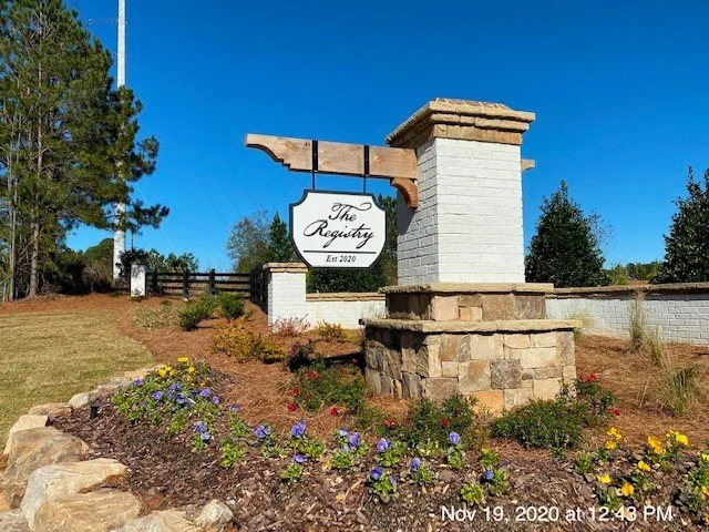 Sign that reads 'The Registry' hanging from a wooden beam, with a brick and stone decorative structure in front, surrounded by flowers and mulch, trees in the background, and a clear blue sky.