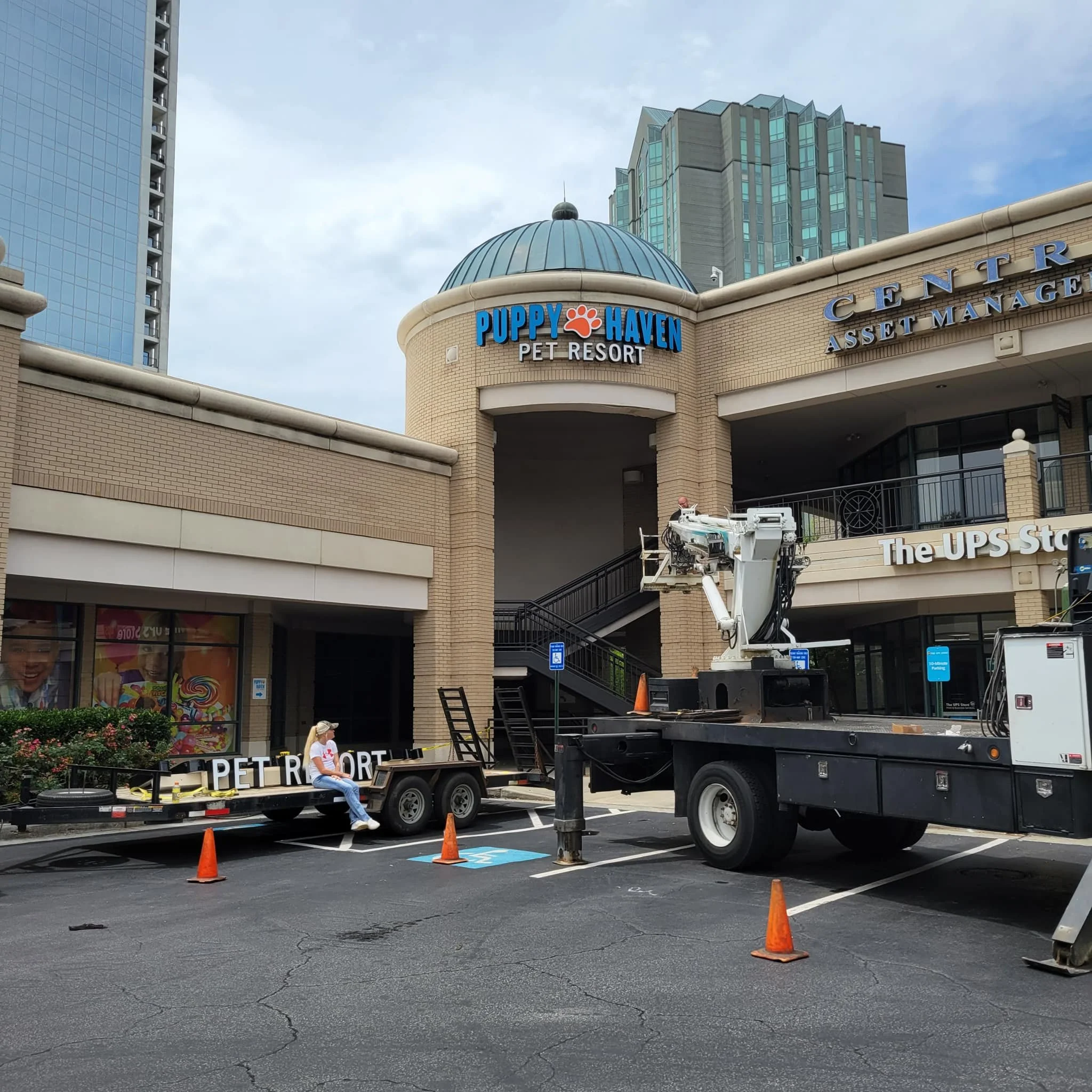 A parking lot outside a shopping center with a sign for Puppy Haven Pet Resort. There is maintenance work being done with a worker sitting on a trailer and a large crane truck parked nearby. The shopping center has brick walls, large windows, and a s