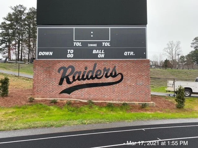 Football scoreboard with Oakland Raiders logo on brick wall beneath it on a sports field.