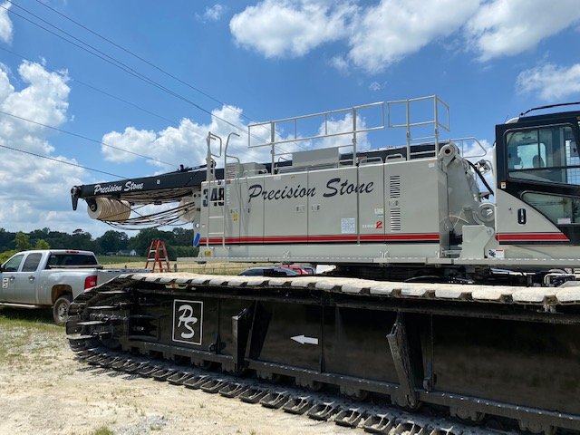A large quarry crane with the words 'Precision Stone' written on the side, mounted on a tracked vehicle, with a pickup truck and blue sky with clouds in the background.