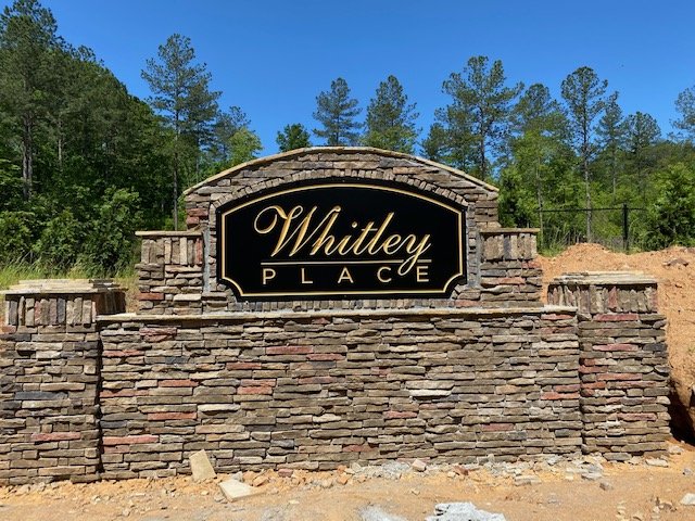 Stone entrance sign for Whitley Place with black background and gold lettering, situated in a semi-rural setting with trees and clear blue sky.