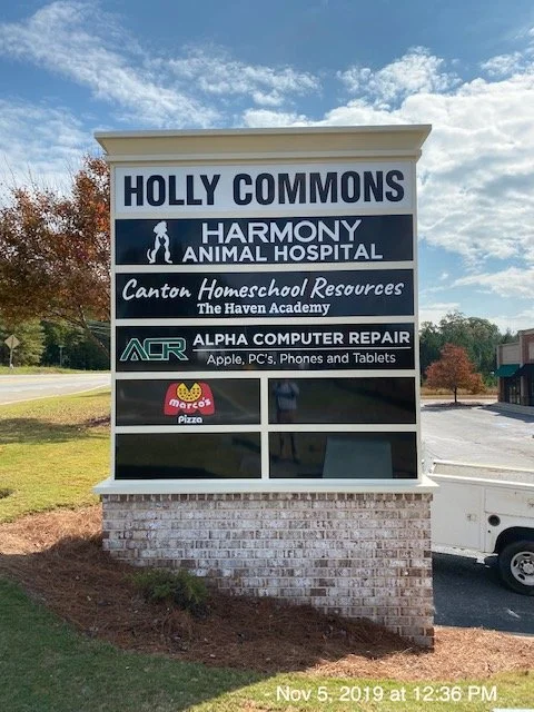 Signboard with multiple business advertisements in Holly Commons, including Harmony Animal Hospital, Canton Homeschool Resources, Alpha Computer Repair, and Marco's Pizza, with a parking lot and trees in the background.