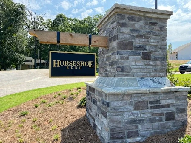Horsehoe Bend sign hanging from a wooden beam attached to a stone pillar at the side of a road.