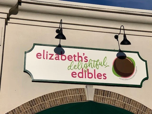Sign for 'Elizabeth's Delightful Edibles' with text and a logo featuring a chocolate circle and a green leaf on a white background.