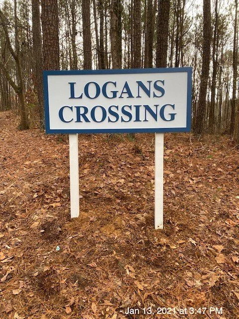 A sign that reads 'Logans Crossing' located in a wooded area with dirt and fallen leaves on the ground.