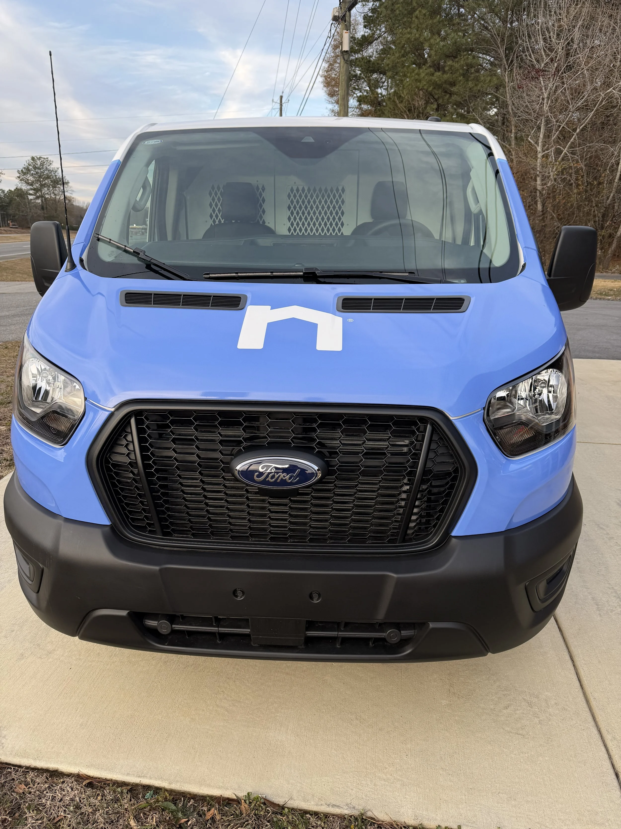 Front view of a blue Ford transit van with a black grille and a white logo on the hood, parked on a concrete surface outside.
