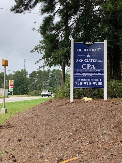 Signboard for Eichelkraut & Associates, Inc., CPA, with contact information, located near a road with a gas station and a few cars passing by, surrounded by trees.