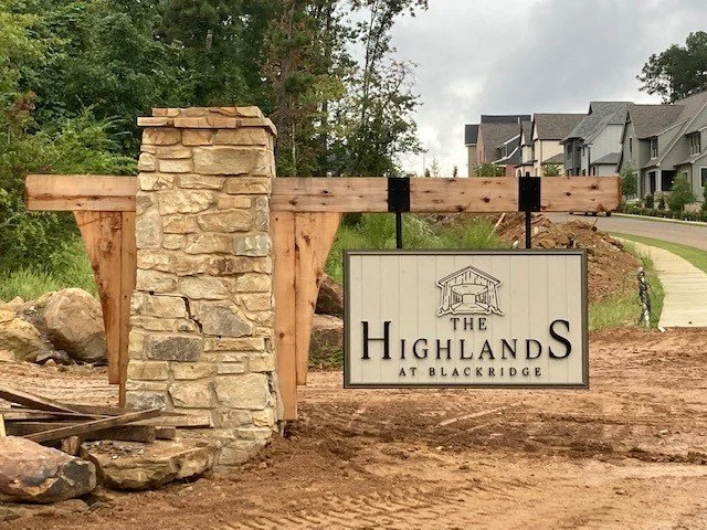 A sign that reads 'The Highlands at Blackridge' attached to a wooden frame, with a stone pillar on the left side, situated in a residential area with new construction in the background.