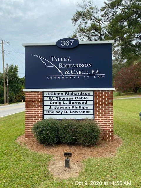Sign for Talley, Richardson & Cable, P.A. law firm with office number 367, listing lawyers J. Glenn Richardson, W. Thomas Cable, Craig L. Burnsed, J. Jayson Phillips, and Chelsley D. Lawrence, situated on a brick base with a shrub in front, in a gras