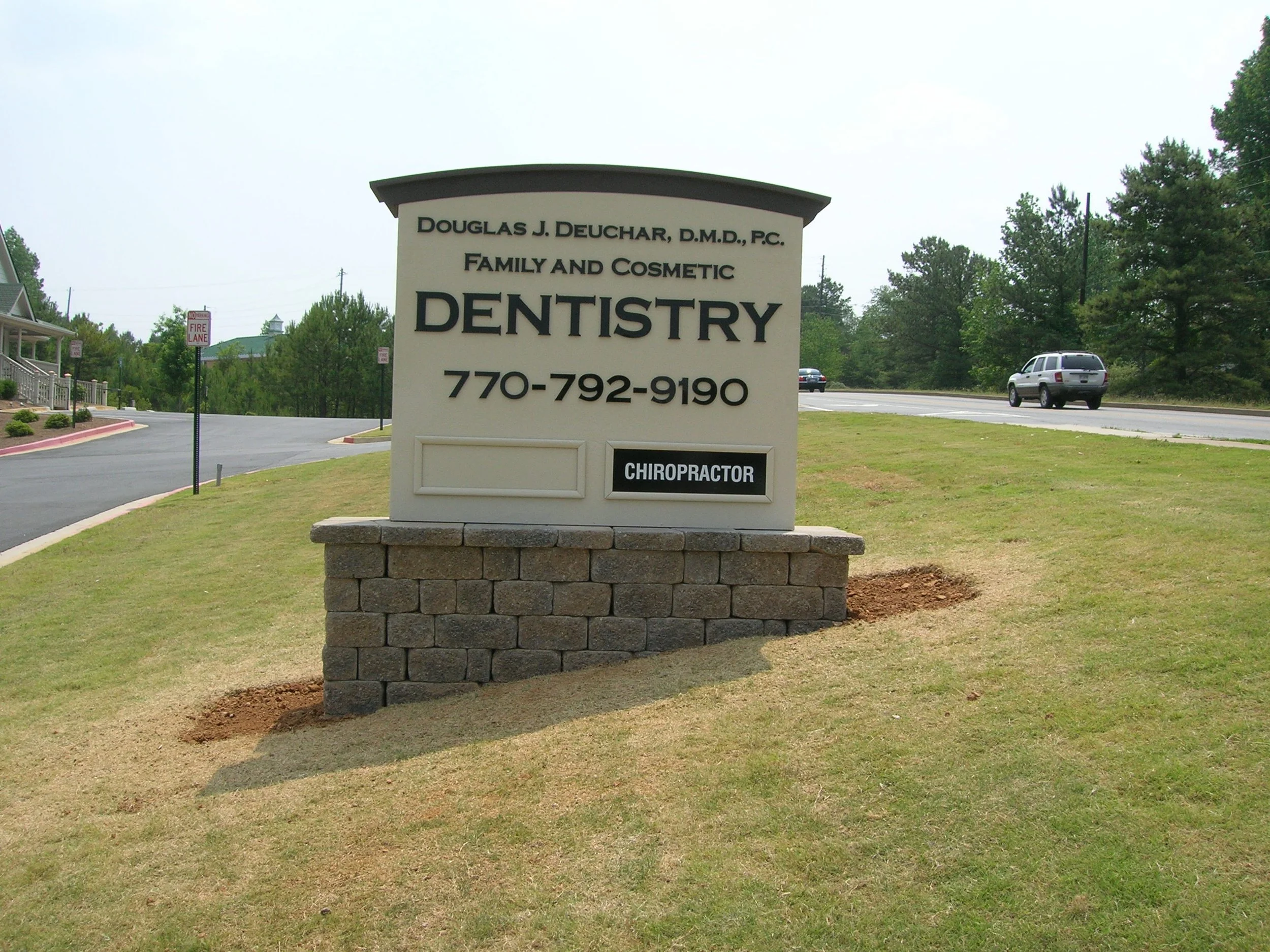 Sign for Douglas J. Deuchar, D.M.D., P.C., Family and Cosmetic Dentistry with phone number 770-792-9190 and a small sign indicating 'Chiropractor' at the bottom, on a grassy area near a parking lot and road with trees in the background.