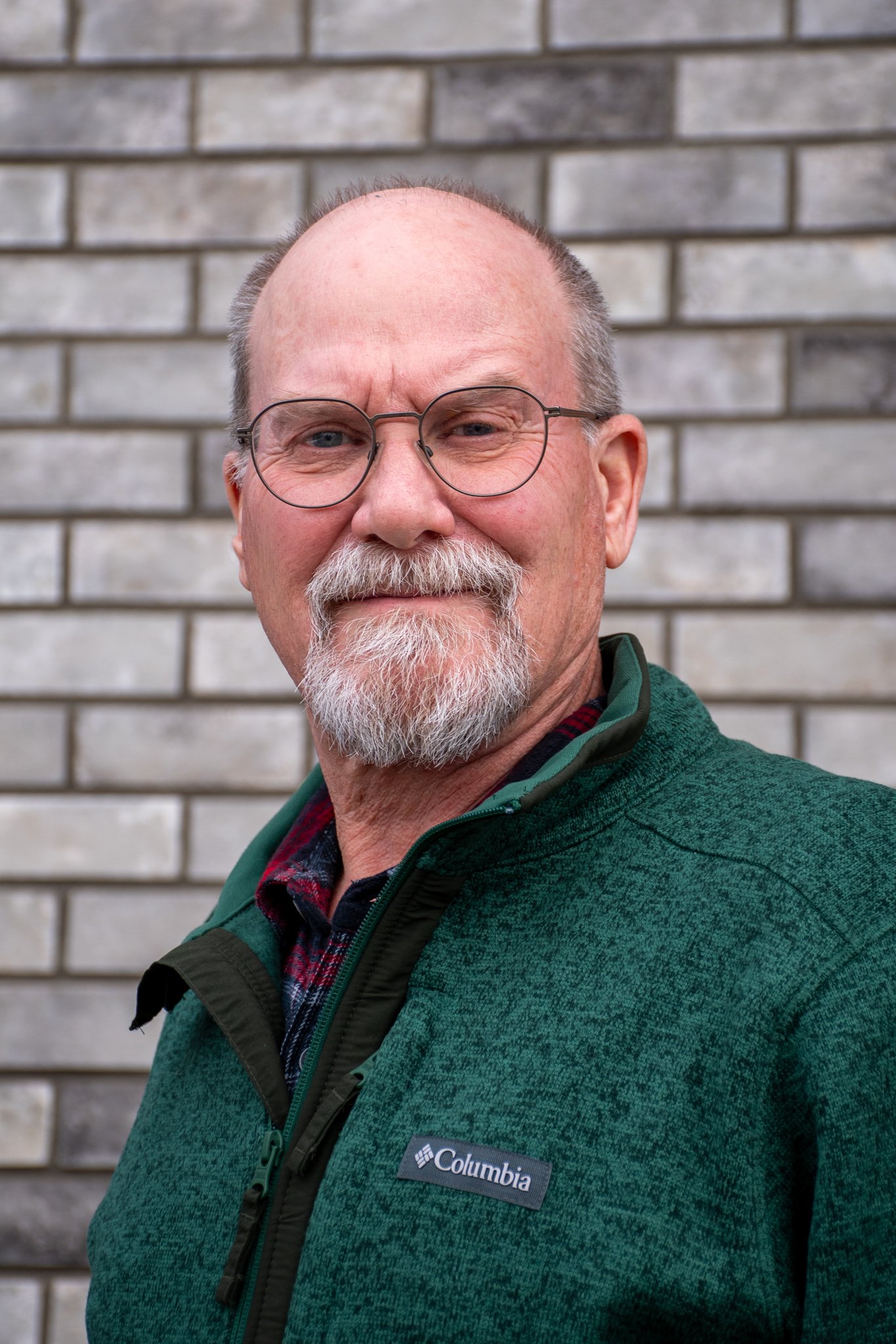 A middle-aged man with glasses and a beard smiling outdoors in front of a brick wall, wearing a green Columbia jacket.