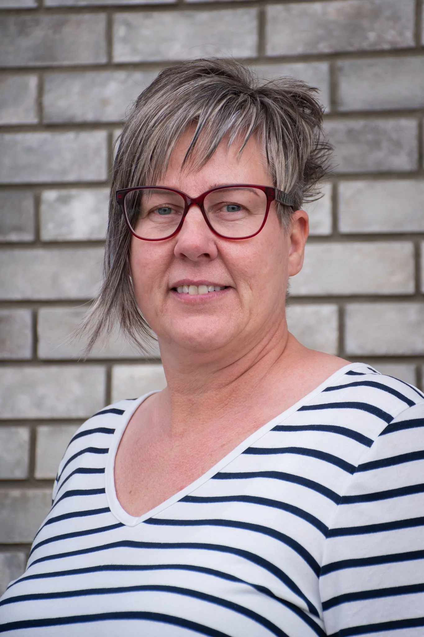 A woman with short, gray hair, wearing red glasses and a navy and white striped shirt, standing in front of a gray brick wall.