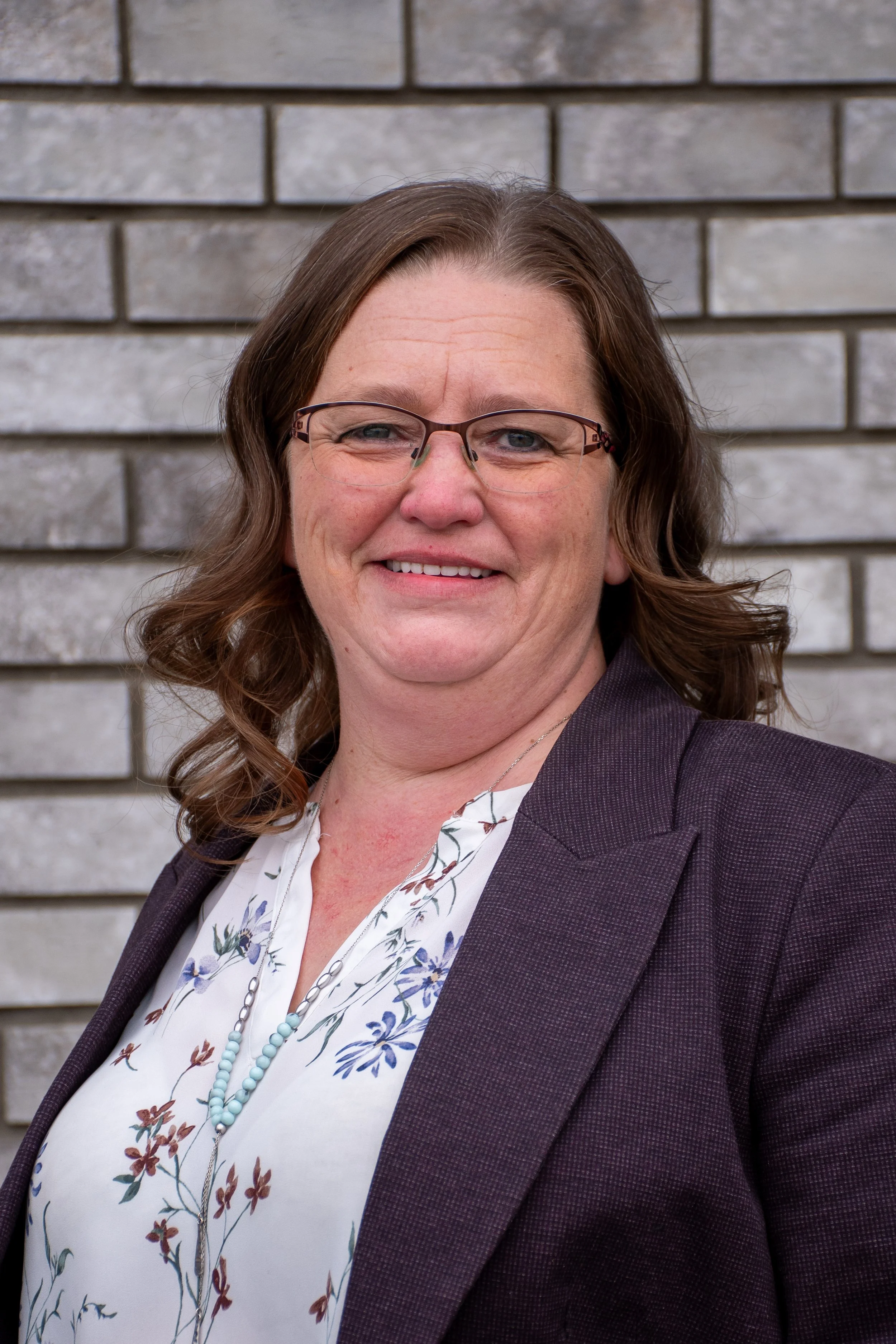 A woman with light skin and brown hair wearing glasses, a floral blouse, a dark blazer, and a beaded necklace, standing in front of a brick wall, smiling at the camera.
