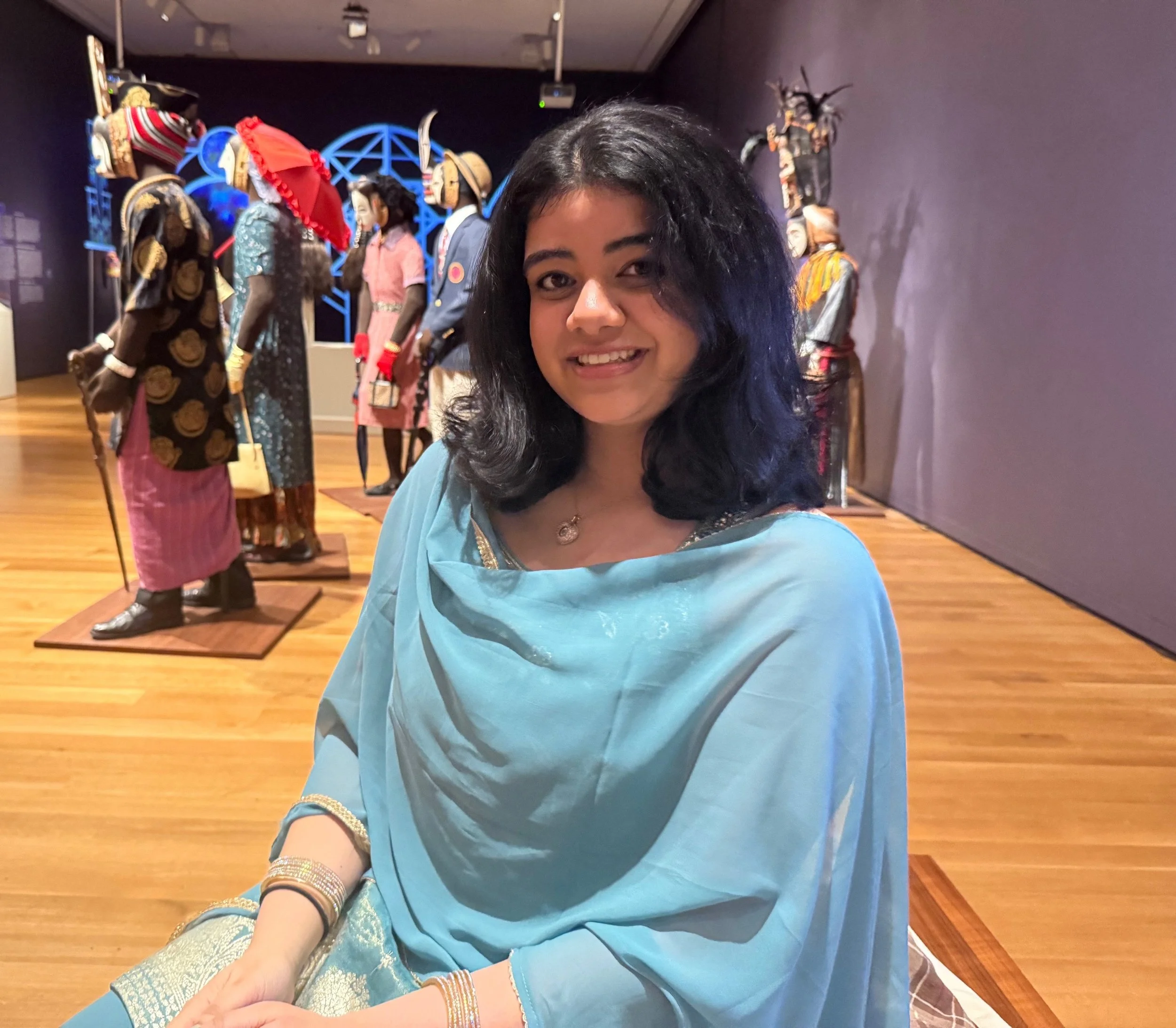 A smiling woman in a light blue outfit sits in front of colorful exhibit displays in a museum-like setting.