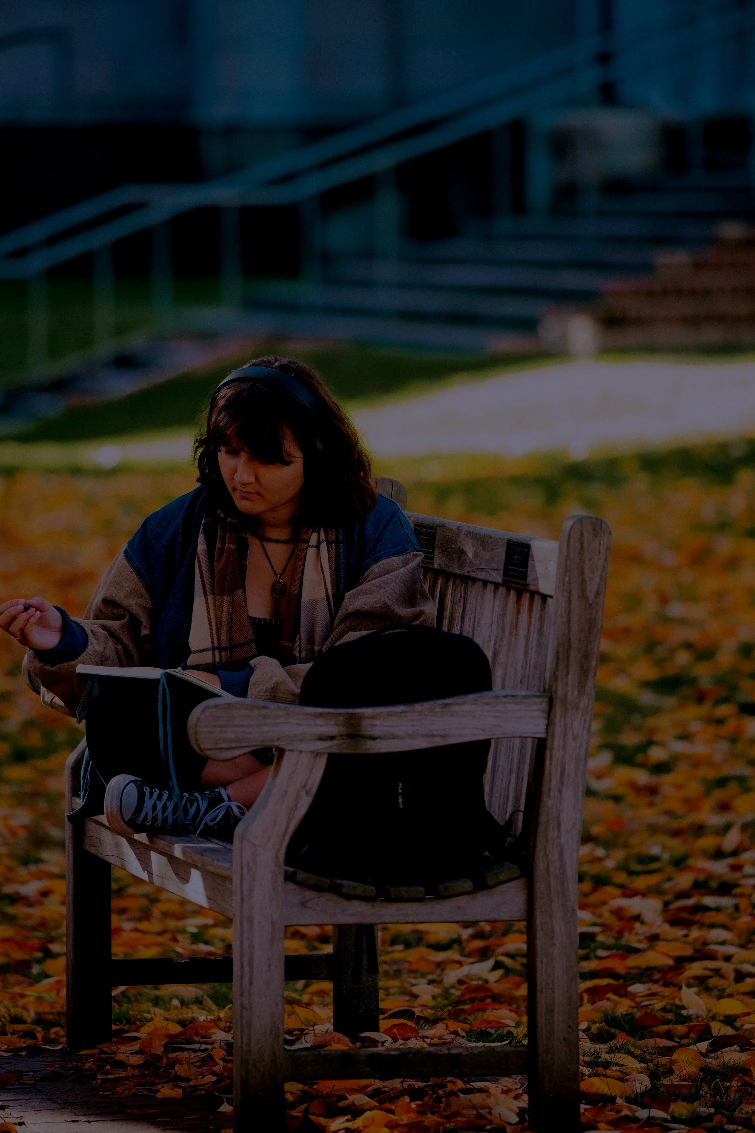 A student reading a book on a wooden bench, with fallen leaves on the ground and a stairway in the background