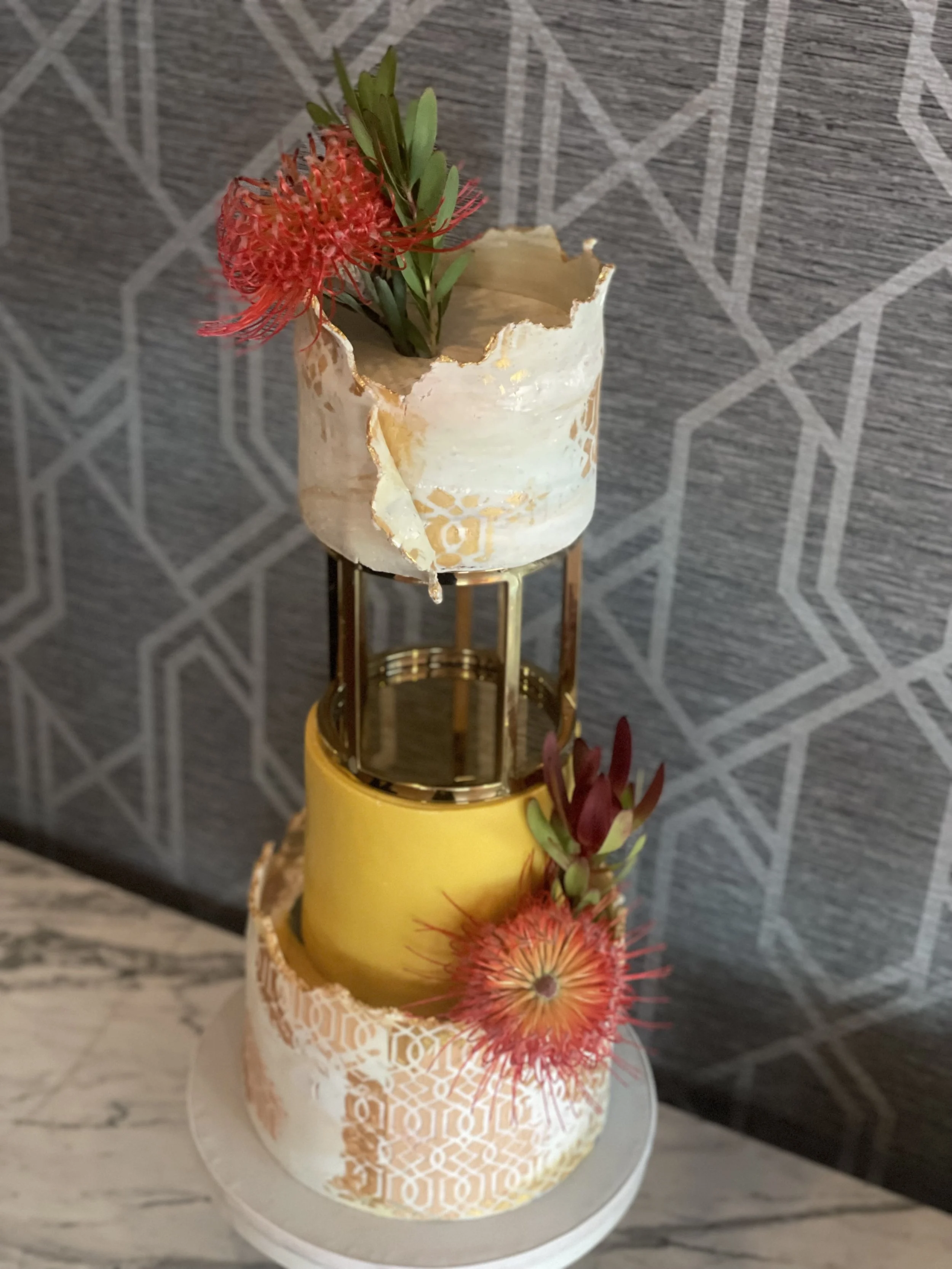 wedding cake with geometric gold and white patterns, topped with flowers and succulents, with a yellow candle in the middle tier.
