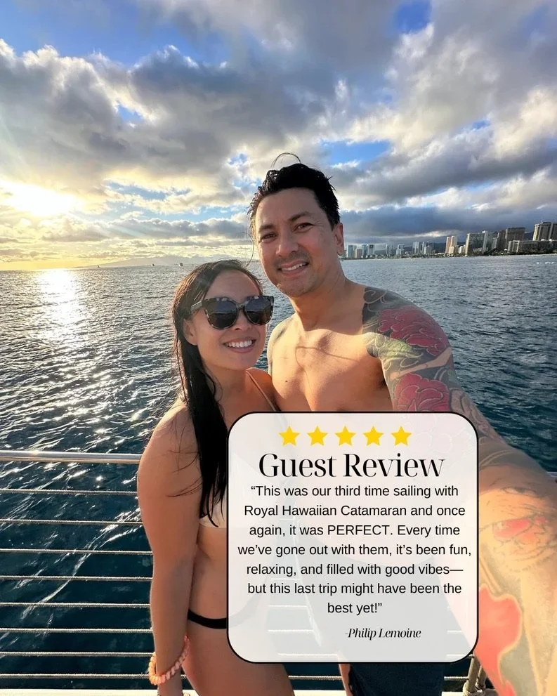 A smiling couple in swimwear taking a selfie on a boat during sunset with a city skyline and ocean in the background.