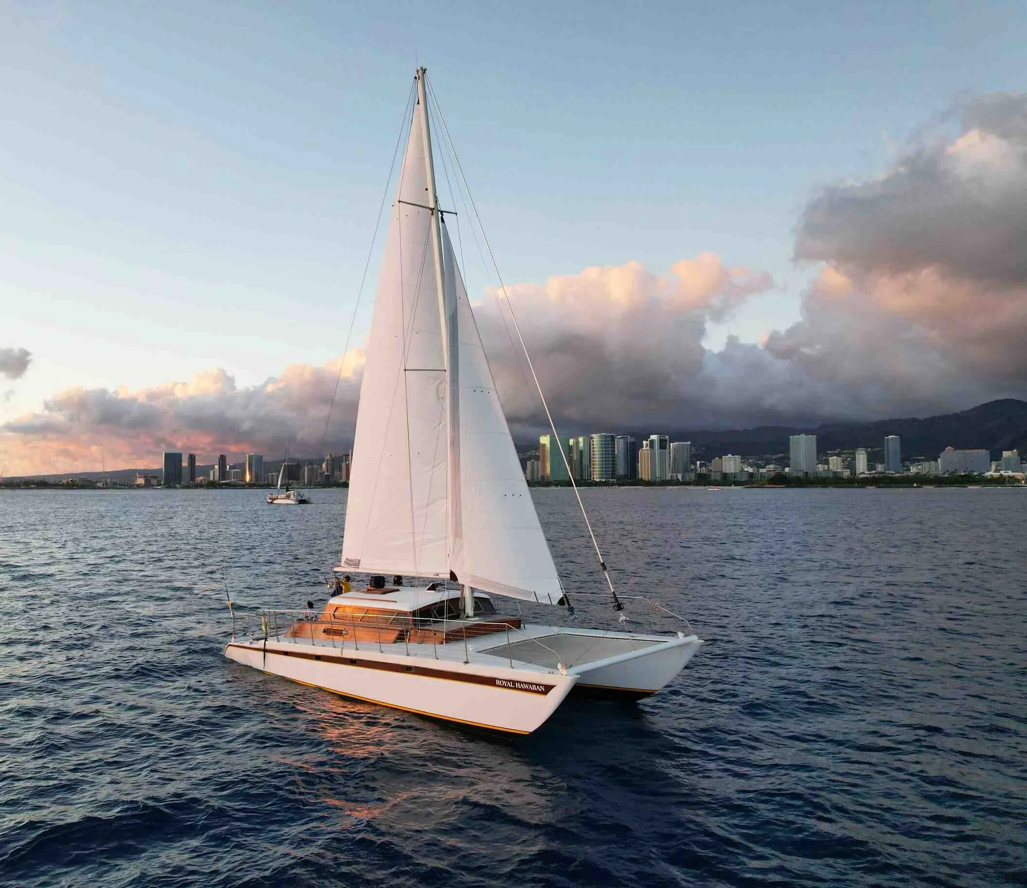 A white sailboat on the water with a city skyline and mountains in the background during sunset.