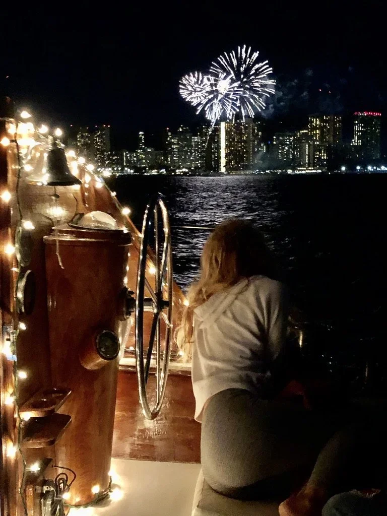 A woman with long blonde hair sitting on a boat deck at night, watching fireworks over a city skyline across the water, with boats and string lights on the boat.