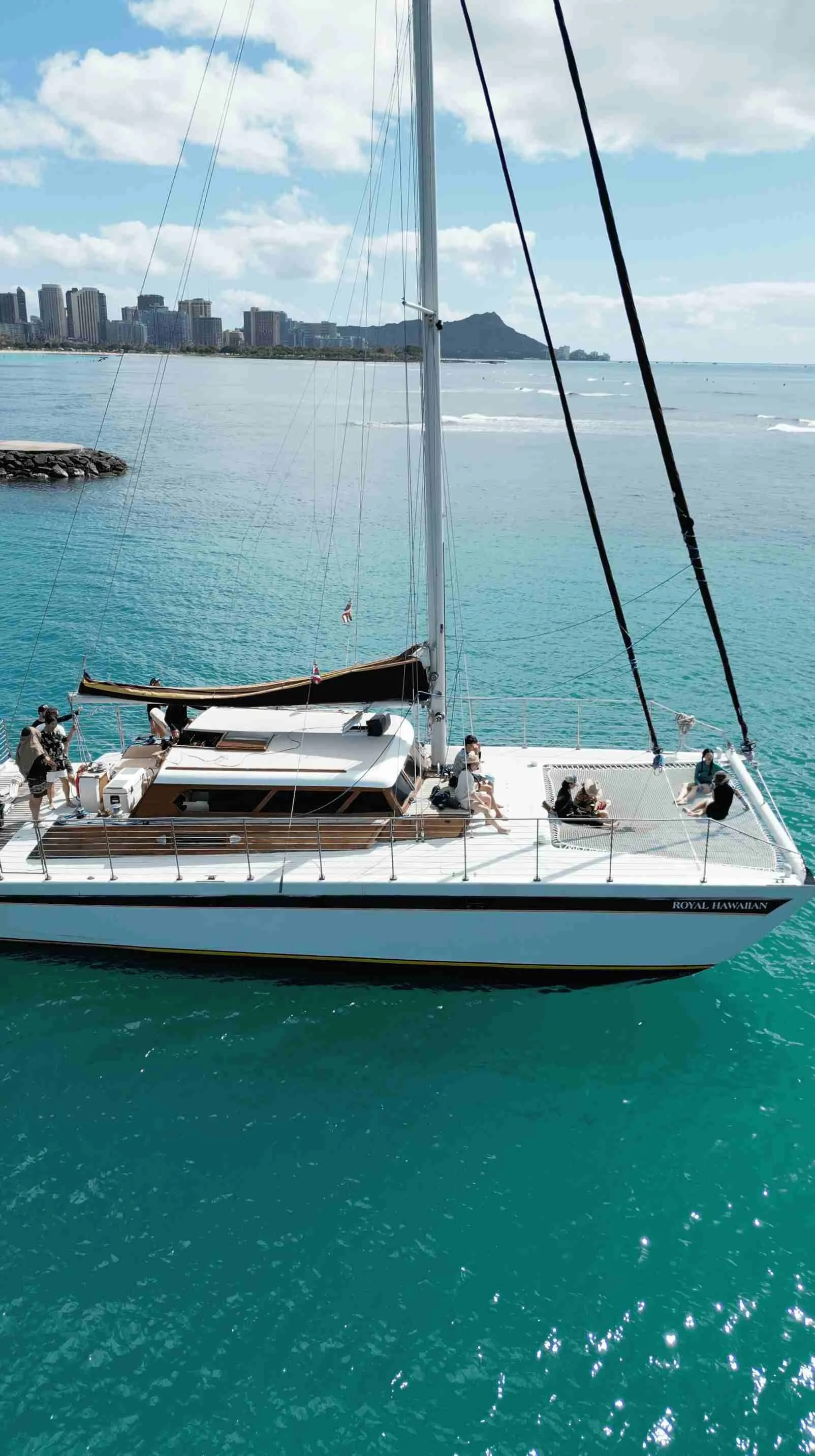 A white sailing boat with people on the deck, floating on turquoise water near a city shoreline with high-rise buildings and mountains in the background.