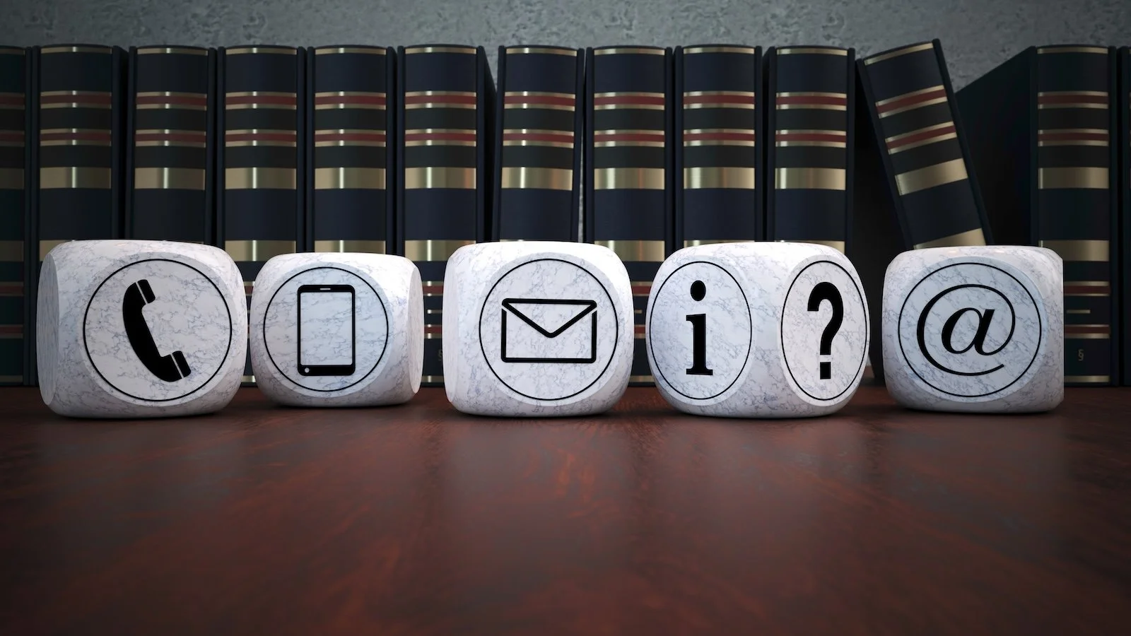 Five white marble blocks with black icons on a wooden surface, representing phone, mobile, email, information, question mark, and at symbol, with a background of black books with gold accents.