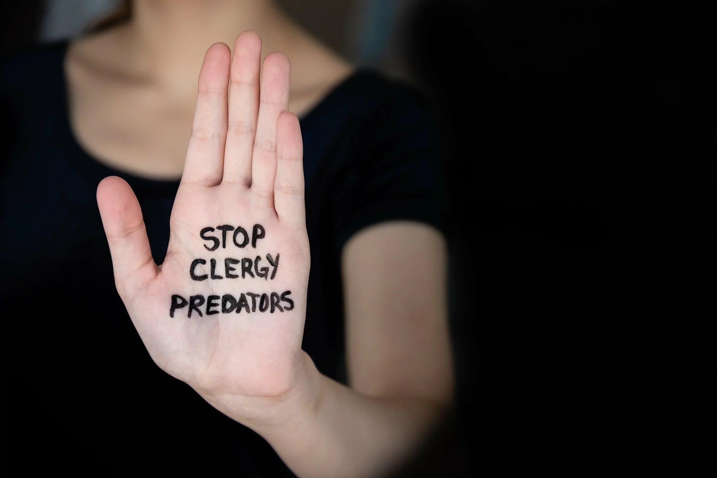 A person's hand with the words 'STOP CLERGY PREDATORS' written on it in black ink, raised in a stop gesture.