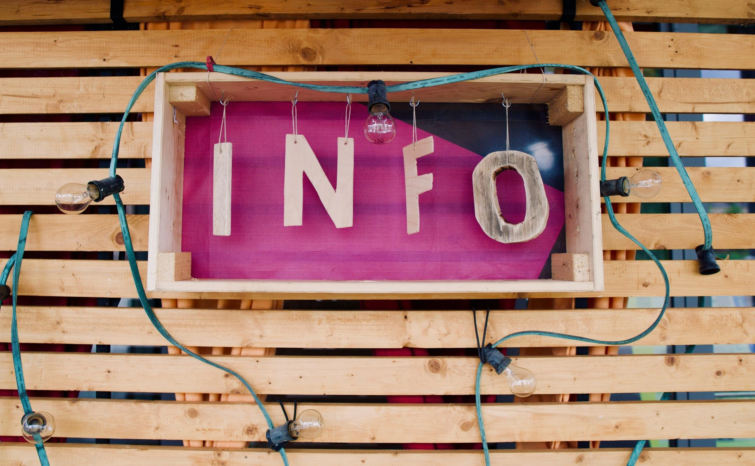 Wooden box with pink background displaying the word 'INFO' with wooden letters, surrounded by string lights, mounted on a wooden slat wall.