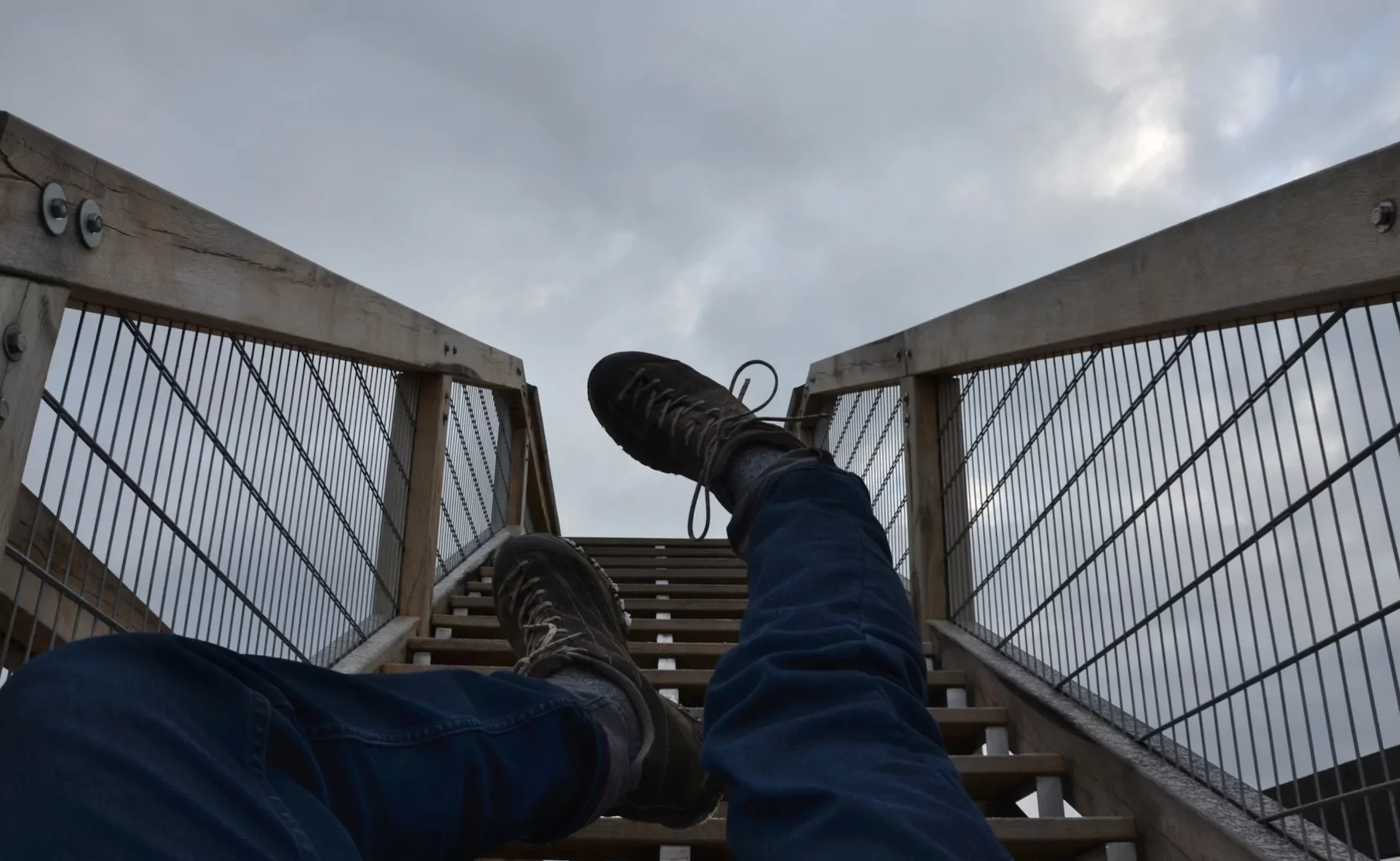 A person sitting on stairs at the top of an outdoor wooden staircase, viewing the cloudy sky, wearing black shoes and dark blue jeans.