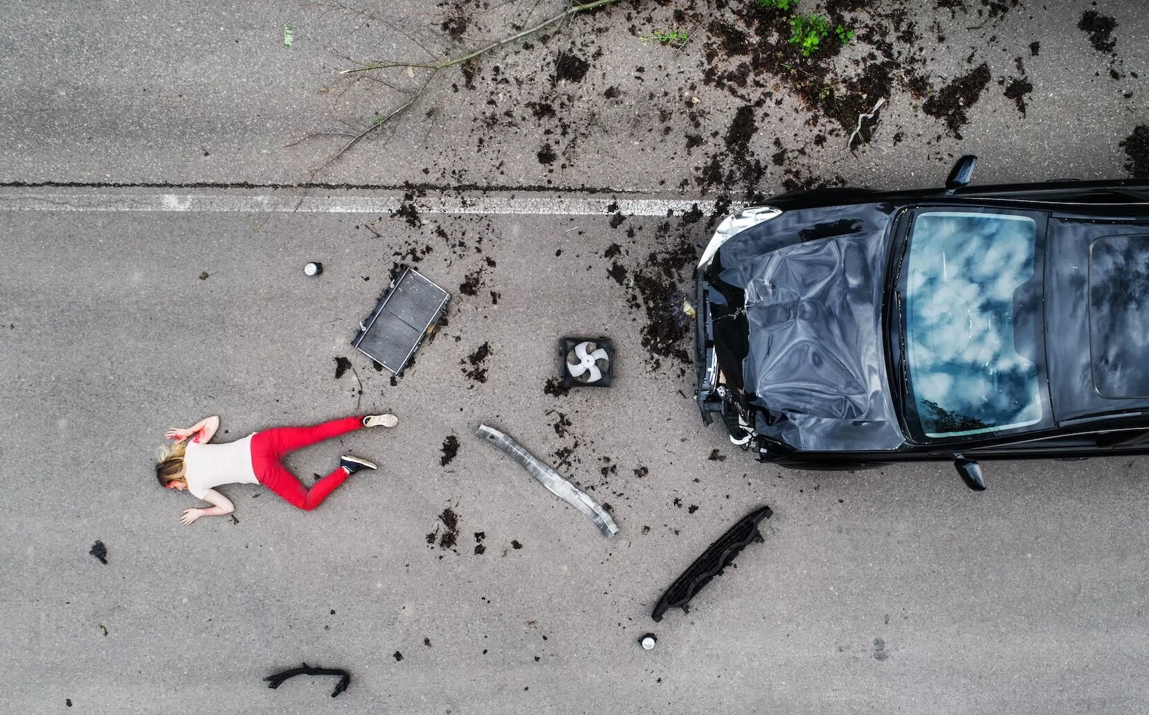 Overhead view of a car accident scene with a damaged black car, scattered debris, and a woman lying on the road.