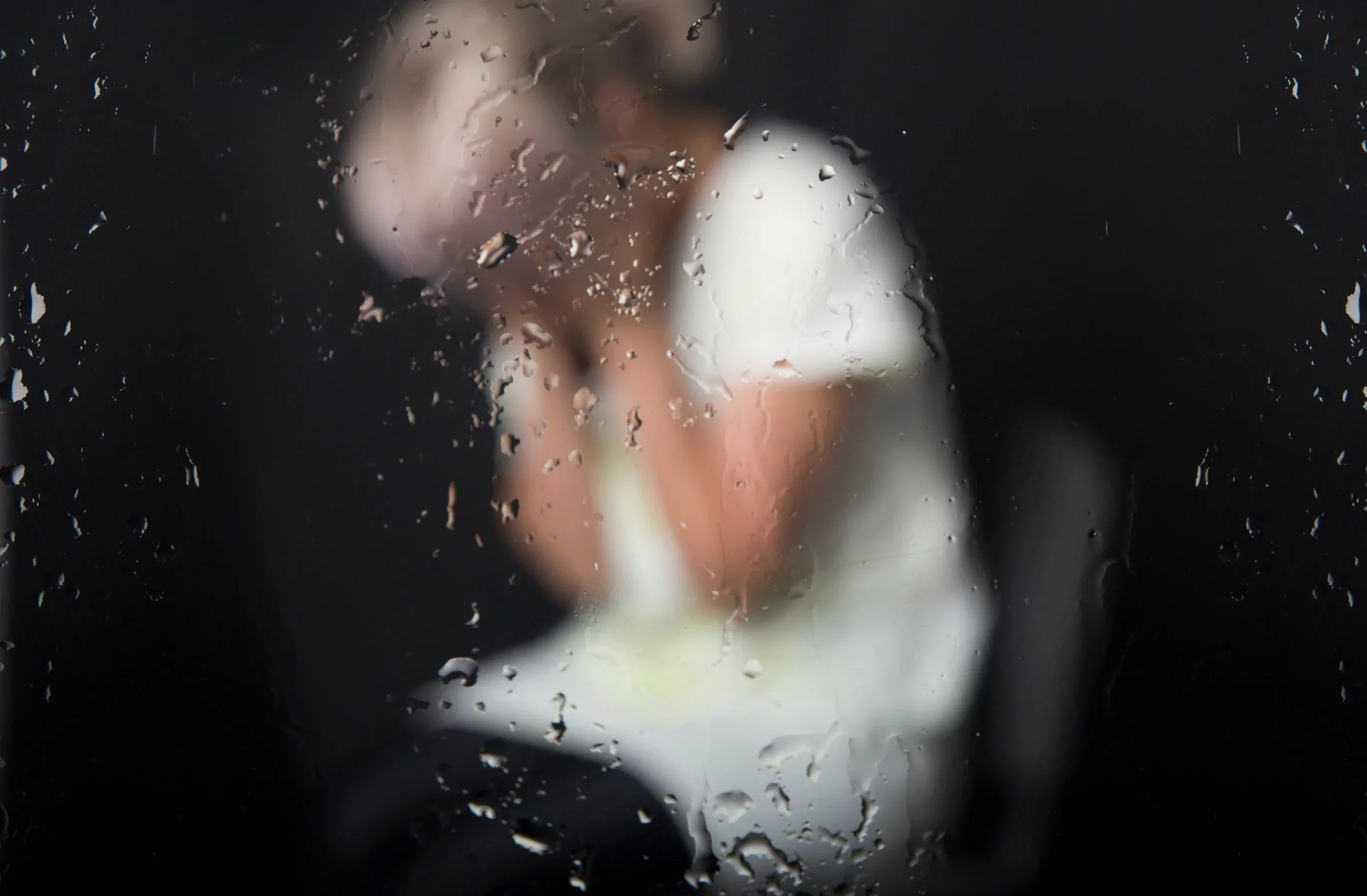 A woman dressed in formal attire is seen through a rain-covered glass window, with water droplets obscuring the view.