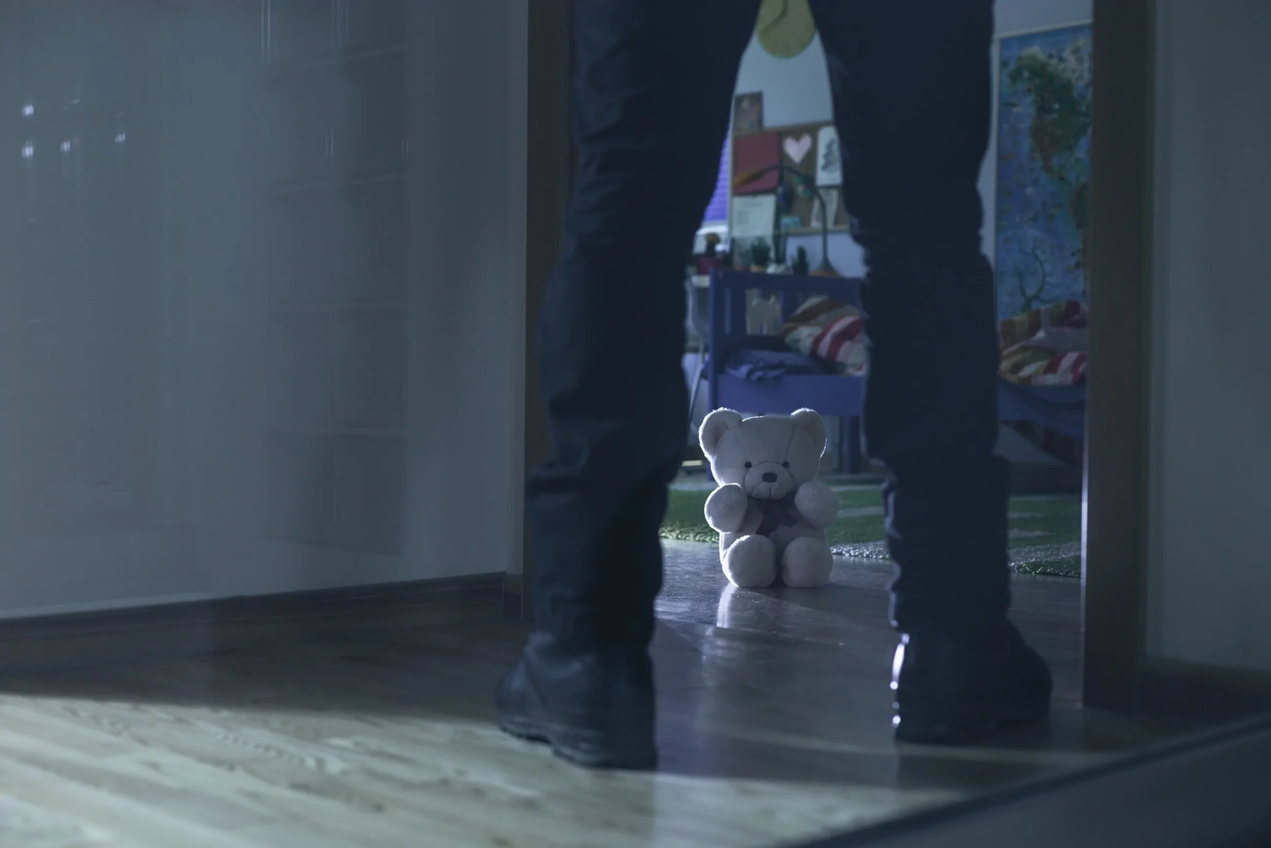 A white teddy bear sitting on a green carpet in front of a window, viewed from behind a person standing in a doorway, with sunlight shining through.