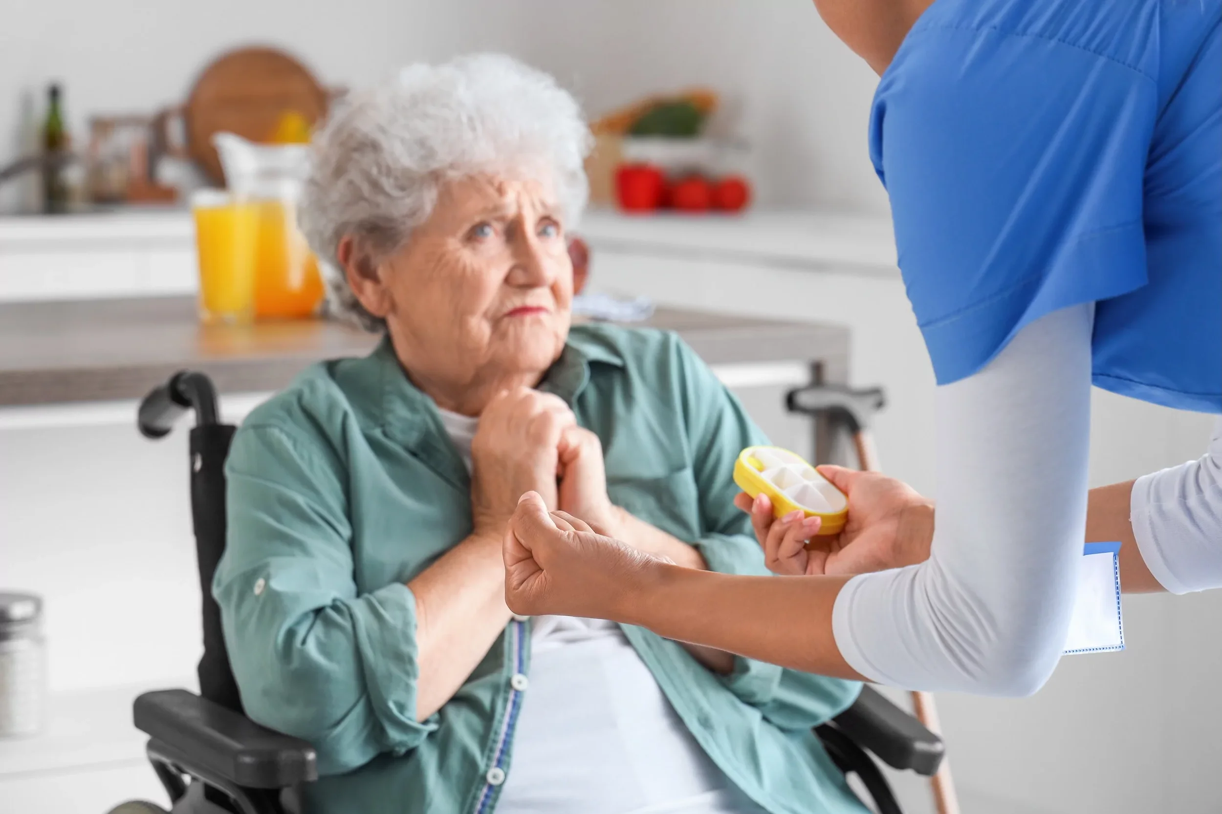 An elderly woman in a wheelchair receives medication from a healthcare worker.