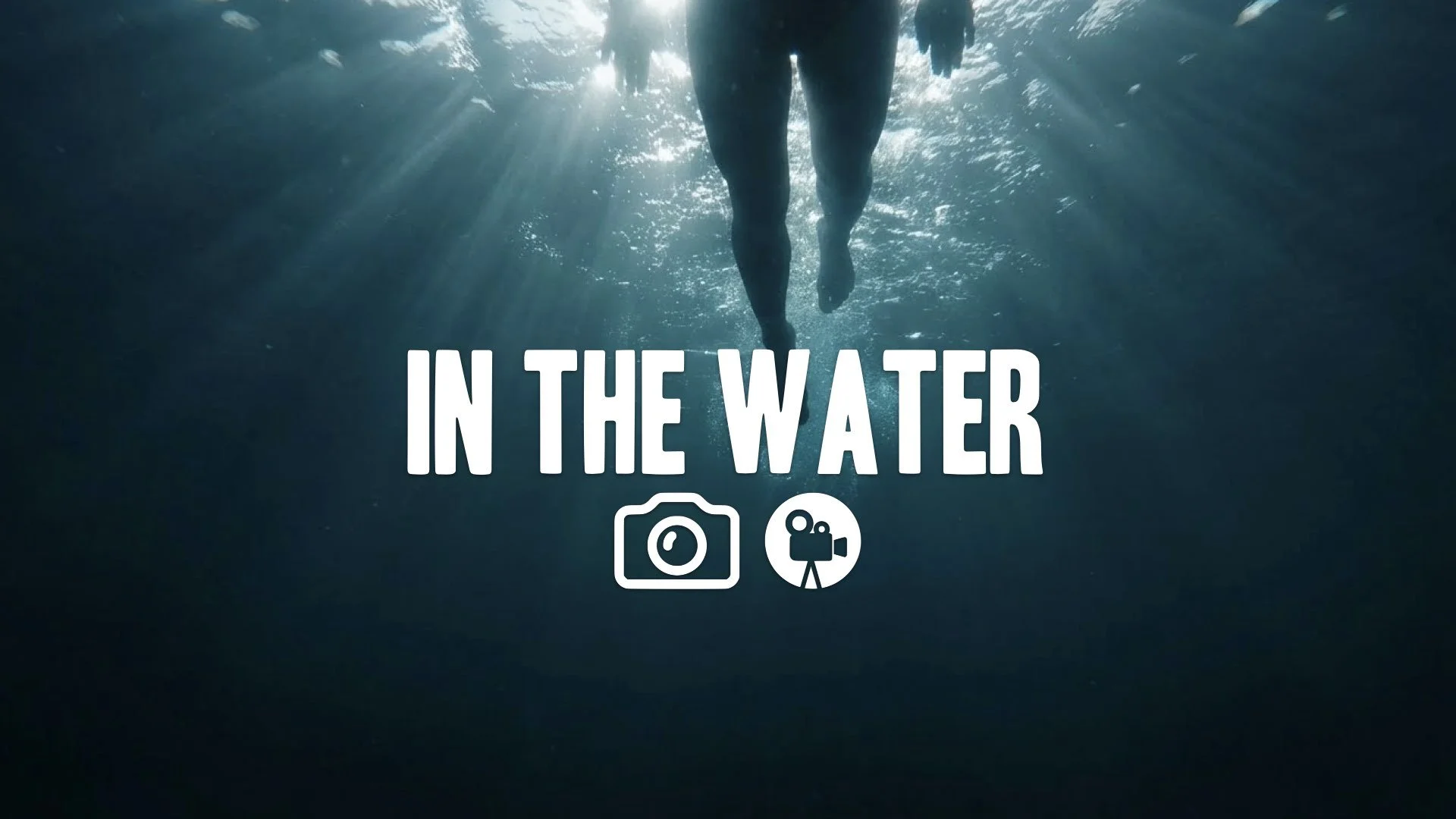 Low-angle POV shot from beneath the water, looking up at a swimmer's legs in the light. Suspenseful, dark blue, no music.