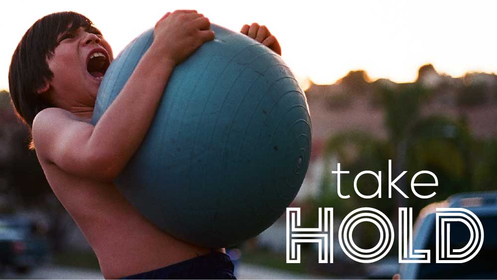 Shirtless boy grasping a playground ball and yelling. 'Take Hold' is written below.