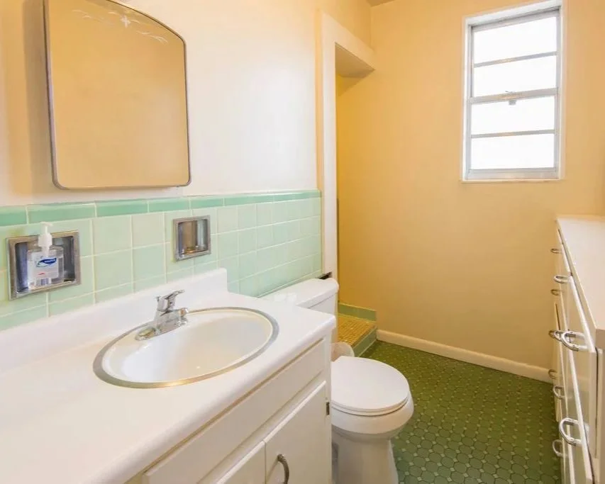Small bathroom with a sink, mirror, toilet, and cabinet. Light green tiled wall behind the sink, yellow wall, and a window with white frame. Green hexagonal tiled floor.