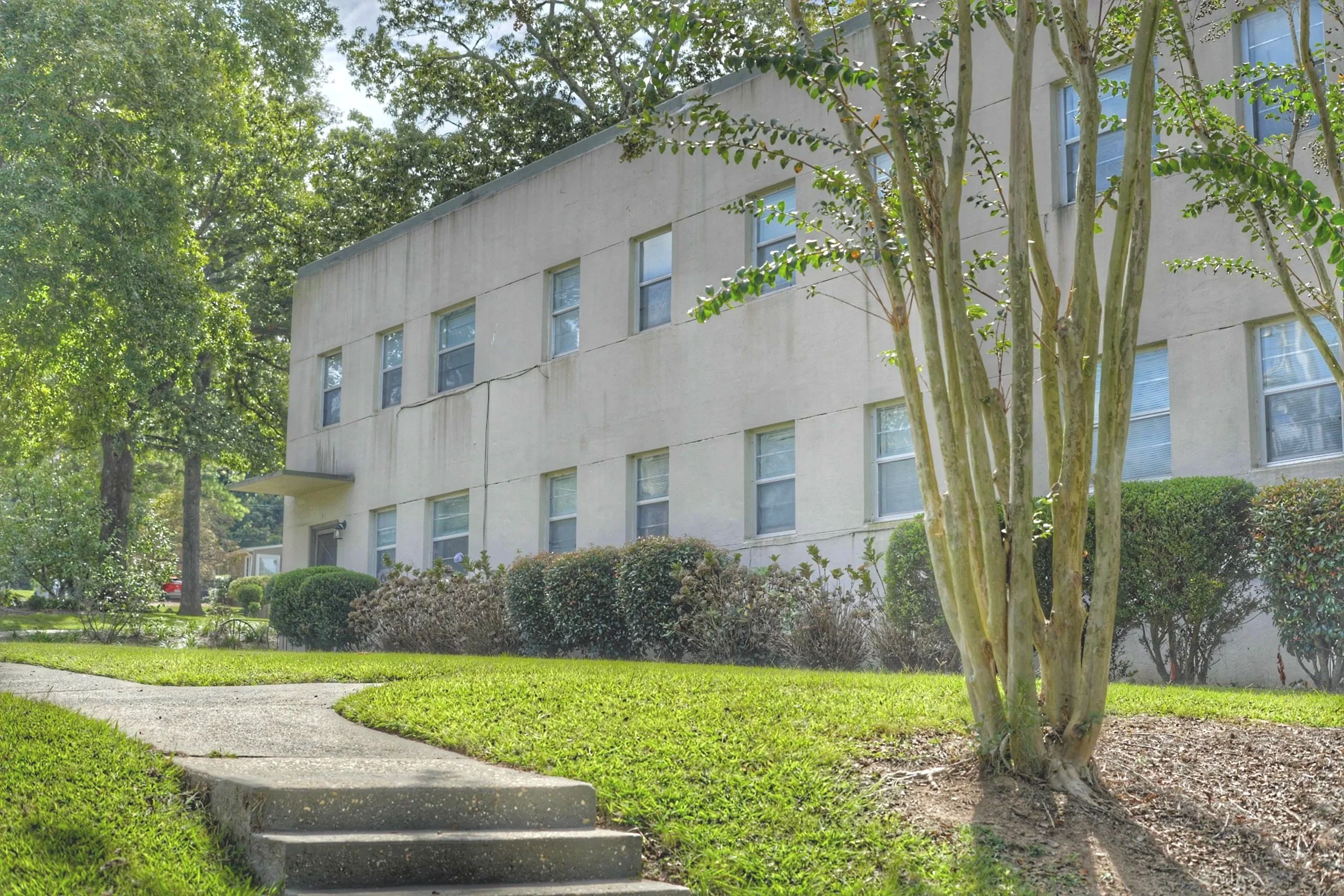 A multi-story residential building with beige exterior walls and multiple windows, surrounded by green trees and bushes, with a sidewalk and concrete steps in the foreground.