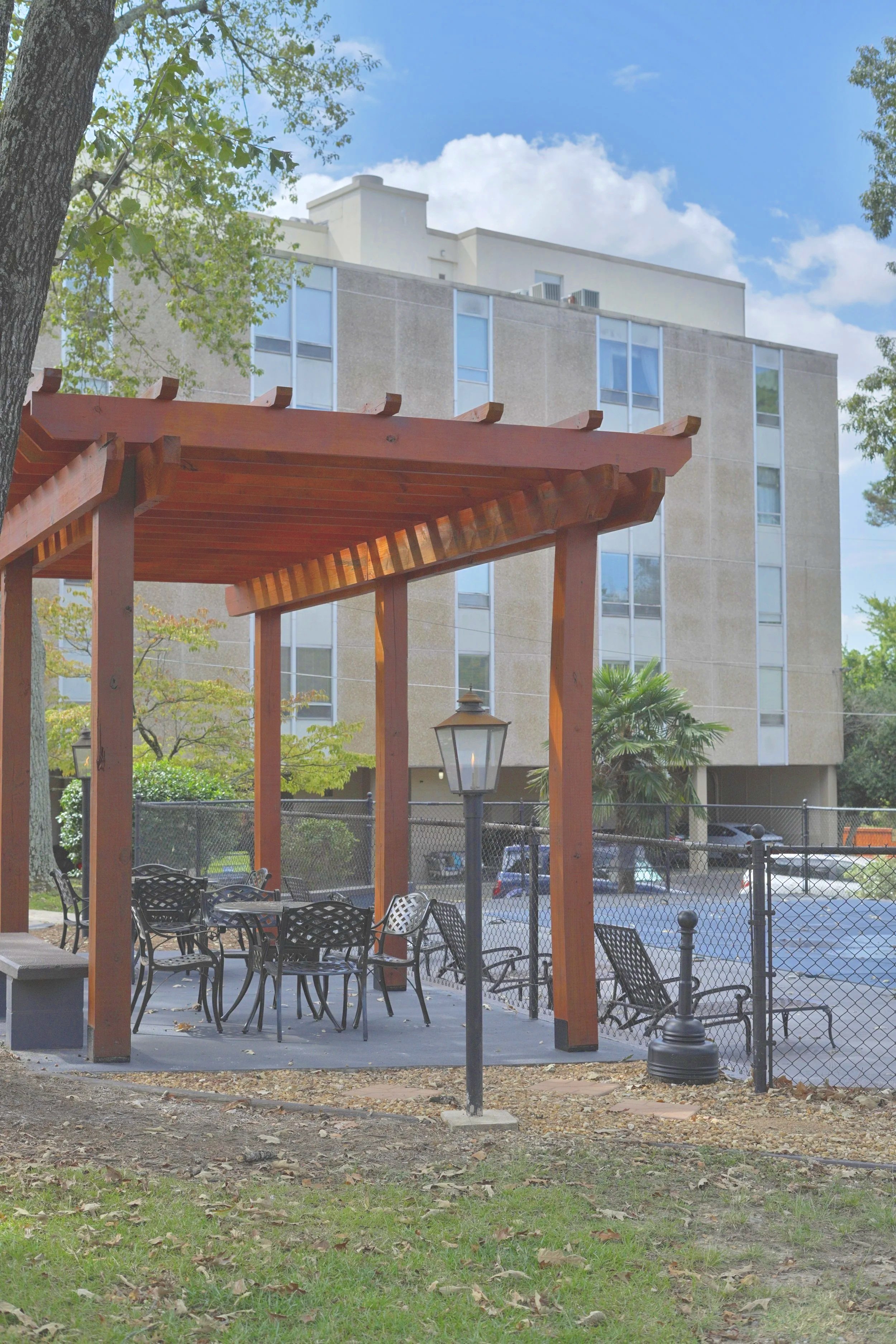 Outdoor seating area with black metal chairs and tables beneath a wooden pergola, surrounded by trees and a residential building in the background.