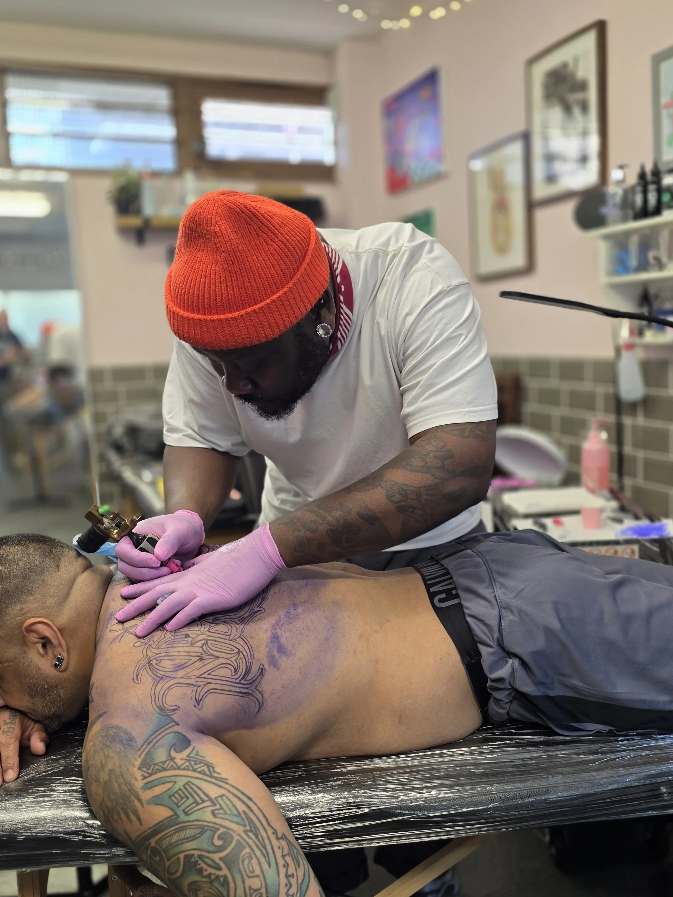 A client with tattoos lying face down on a tattoo table, receiving a tattoo from Lord at The good Fight tattoo in Camden London. The artist is wearing a red beanie, earrings, and pink gloves, focusing on the tattooing process.