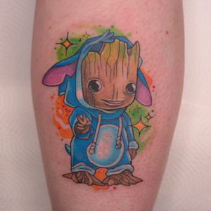 Tattoo of Baby Groot from Guardians of the Galaxy, wearing a blue hoodie, making a peace sign with his fingers, surrounded by colorful stars.