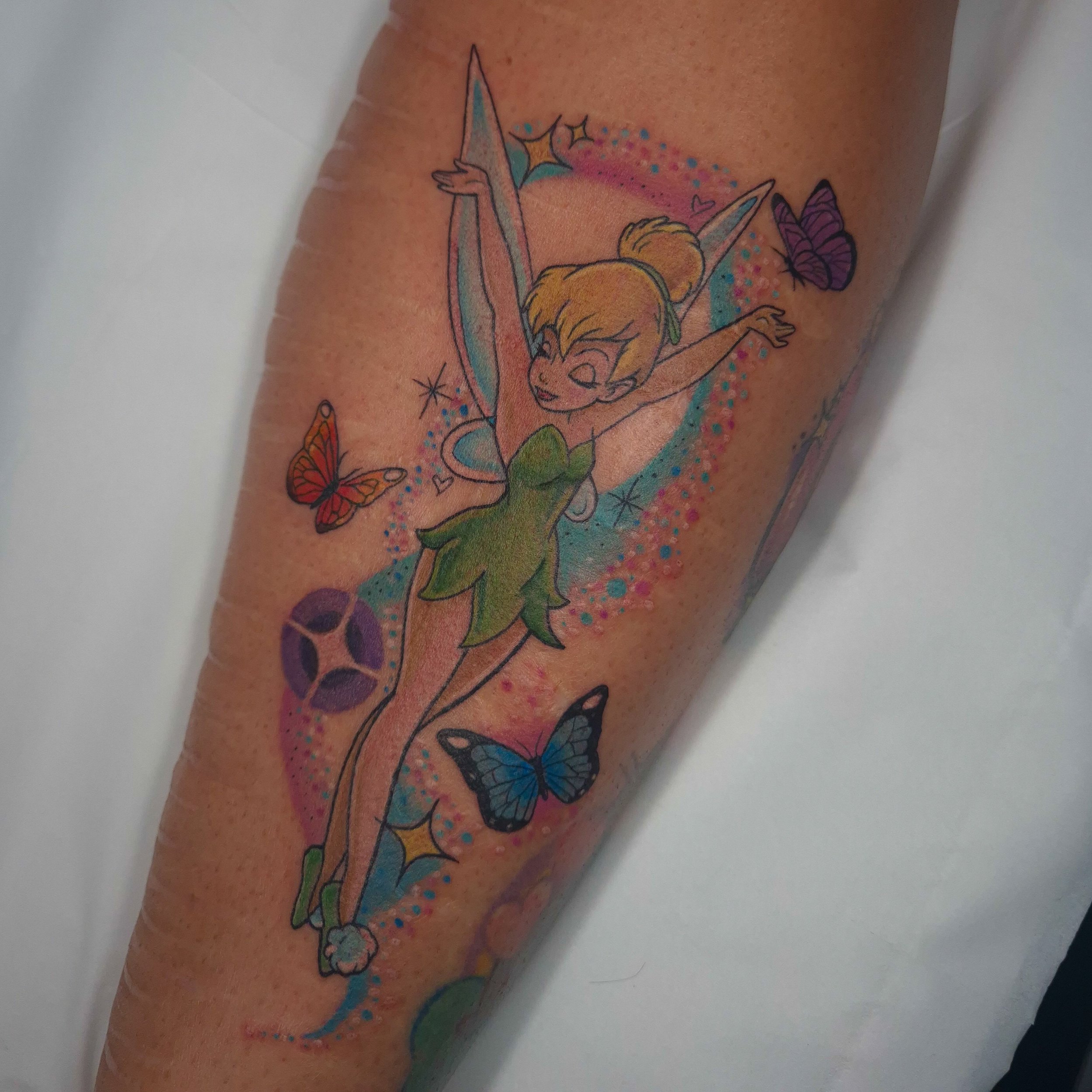 Colorful tattoo of Tinkerbell with butterfly wings, with a pink and blue background and surrounded by butterflies on an arm. Done by Belle at The Good Fight tattoo in Camden London
