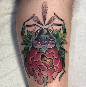 Tattoo of a spider with a body resembling a beetle, surrounded by green leaves and a large red flower.