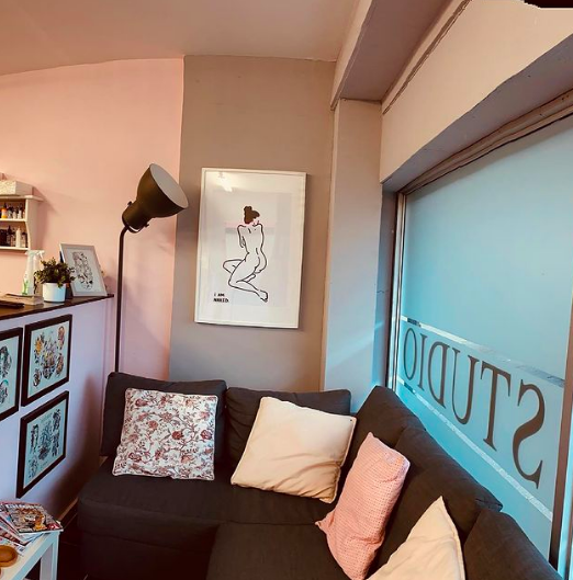 View of a cozy waiting room at The Good Fight tattoo in Camden London with a dark gray couch, decorative pillows, a framed artwork of a kneeling woman on the wall, a tall floor lamp, a window with frosted glass labeled 'STUDIO', and books on a shelf.