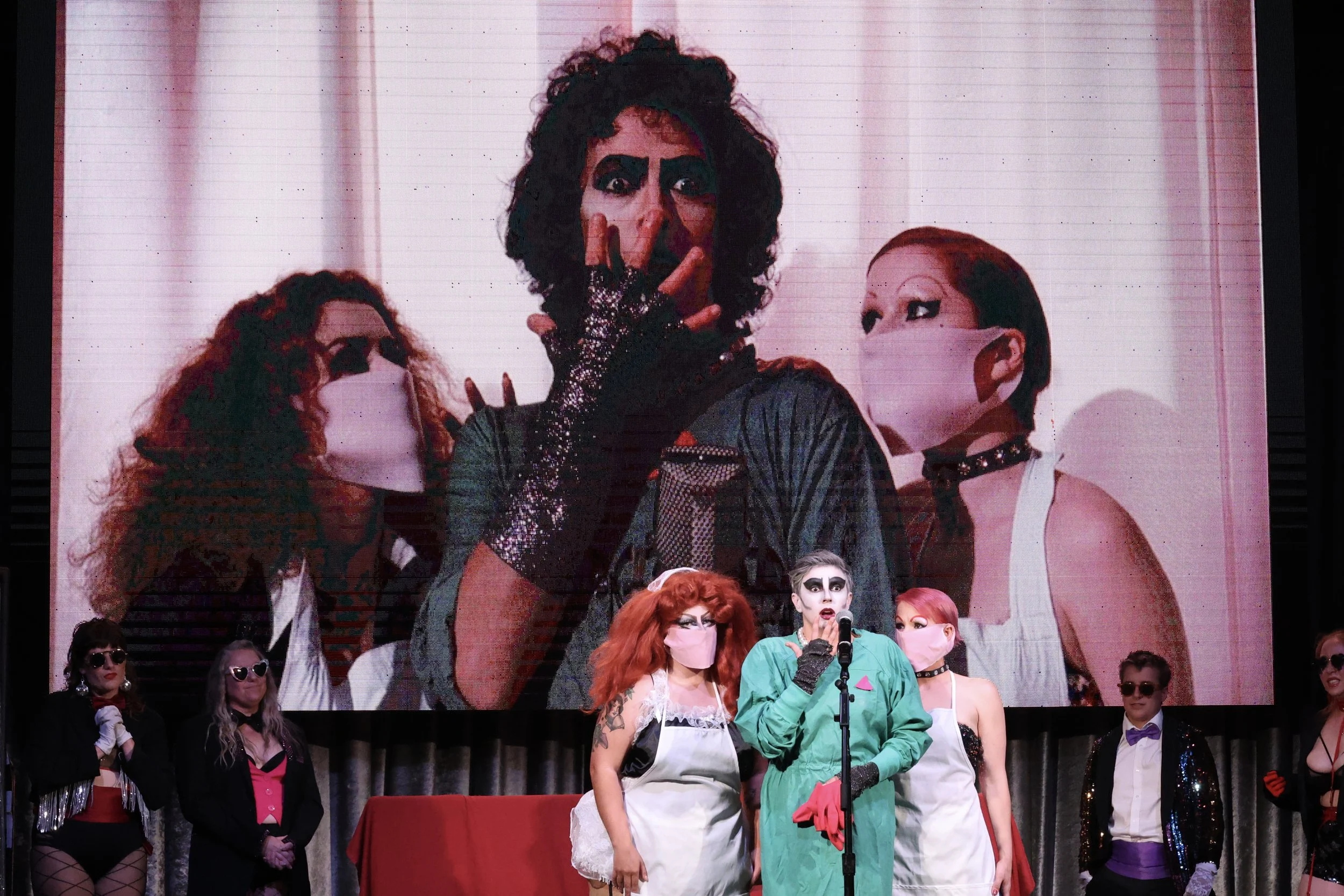 A large screen displays a person with dark curly hair and dramatic makeup posing with fingers in a V shape over their face, surrounded by a group of people all wearing face masks, some with colorful hair and costumes, at a convention or event.