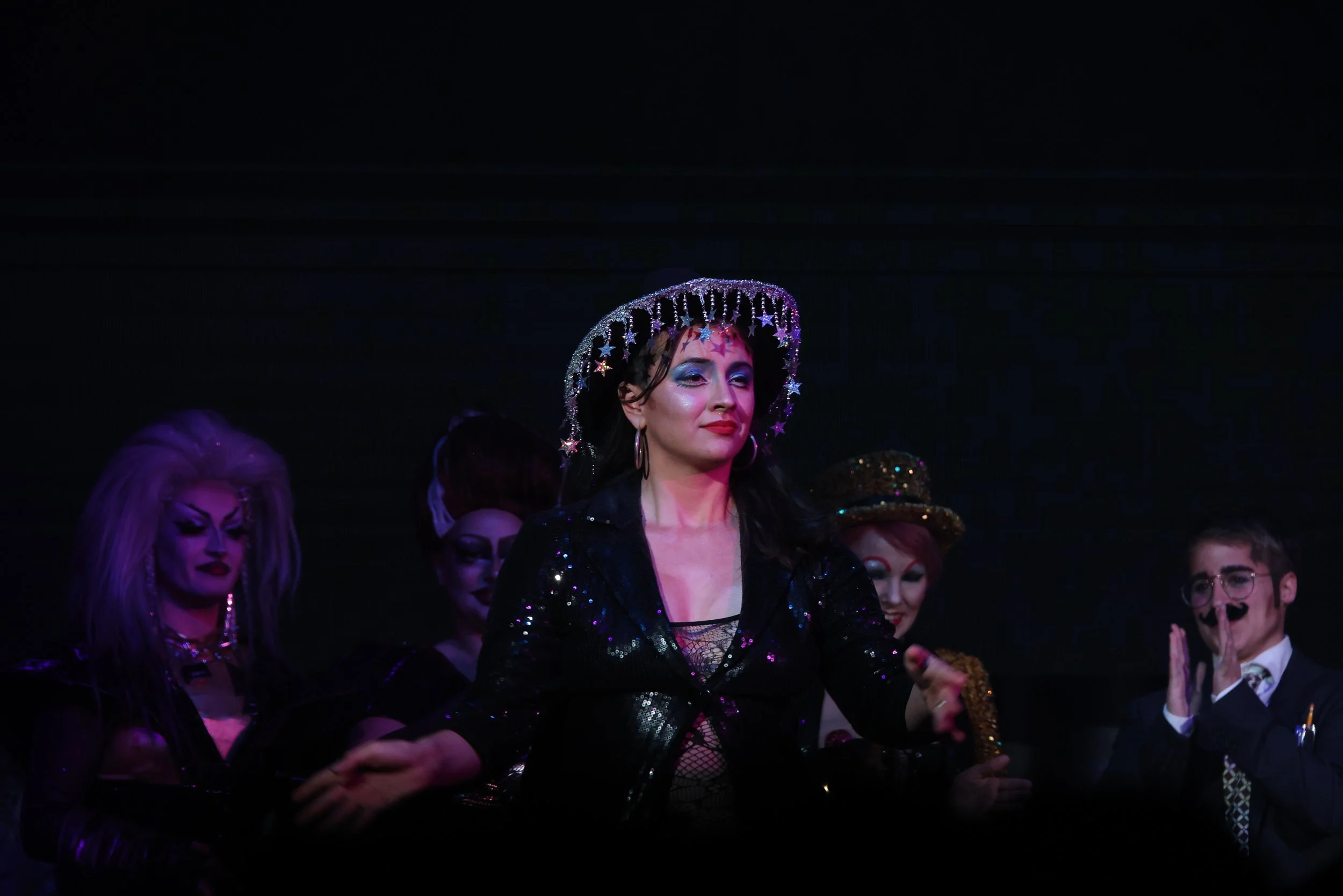 Linnea wearing a glittery black outfit and star-decorated cowboy hat, standing among a group of people with colorful makeup and costumes on a stage.