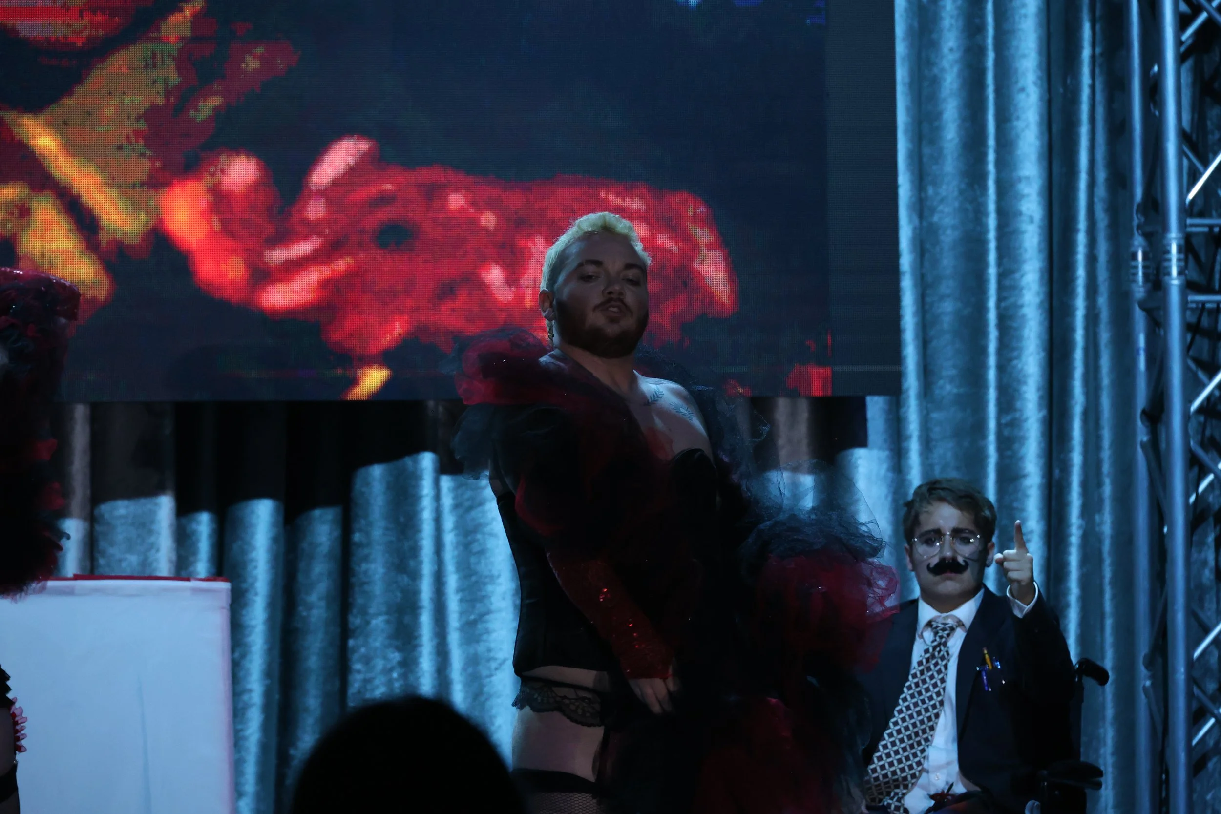 Person with platinum blonde hair and beard standing on stage with a digital background and people in costume, including a person with a mustache and glasses giving a thumbs up.
