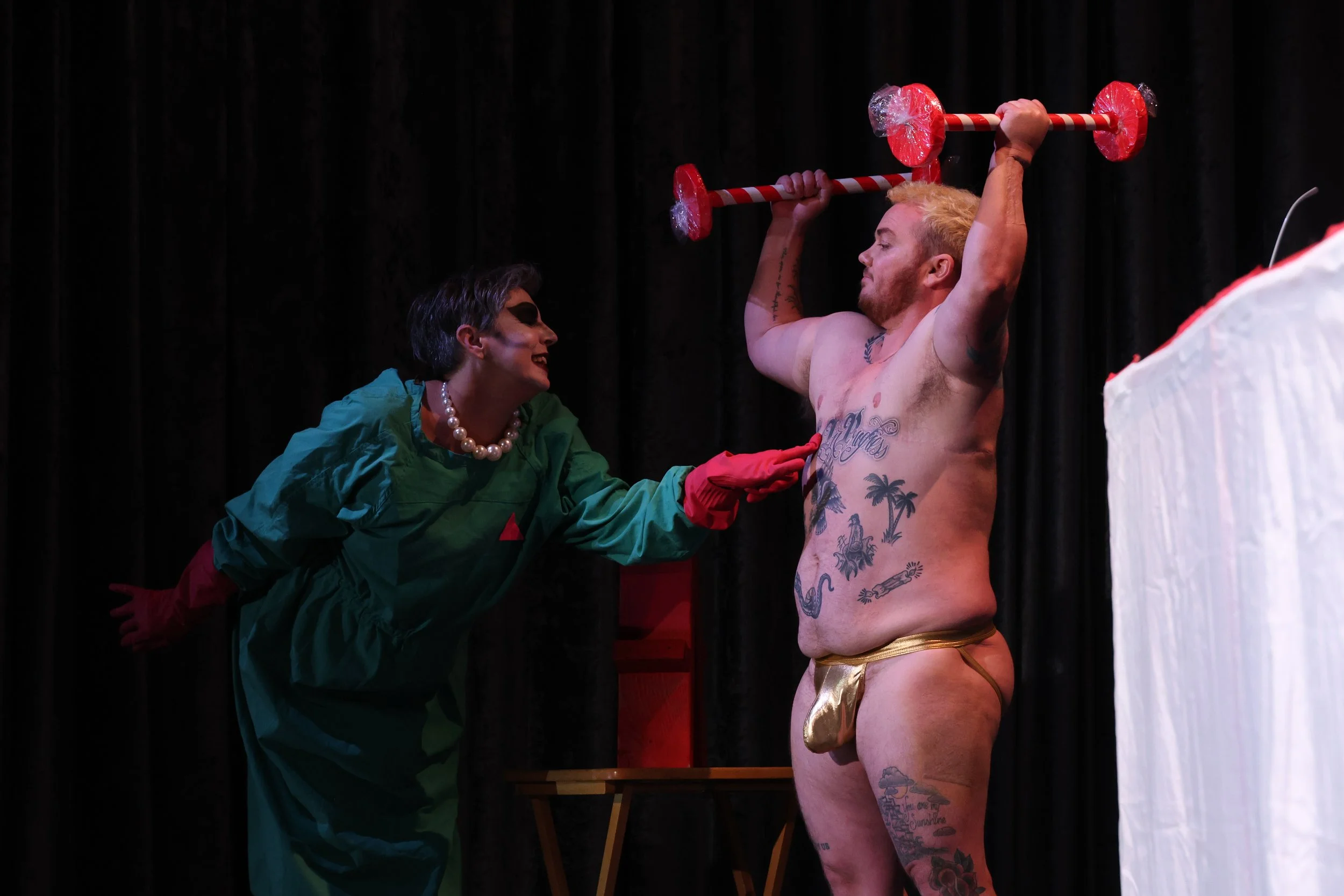 A woman dressed in a green jacket, red gloves, and pearl necklace is looking at a shirtless man holding a large candy cane-shaped barbell above his head, with a black curtain in the background.