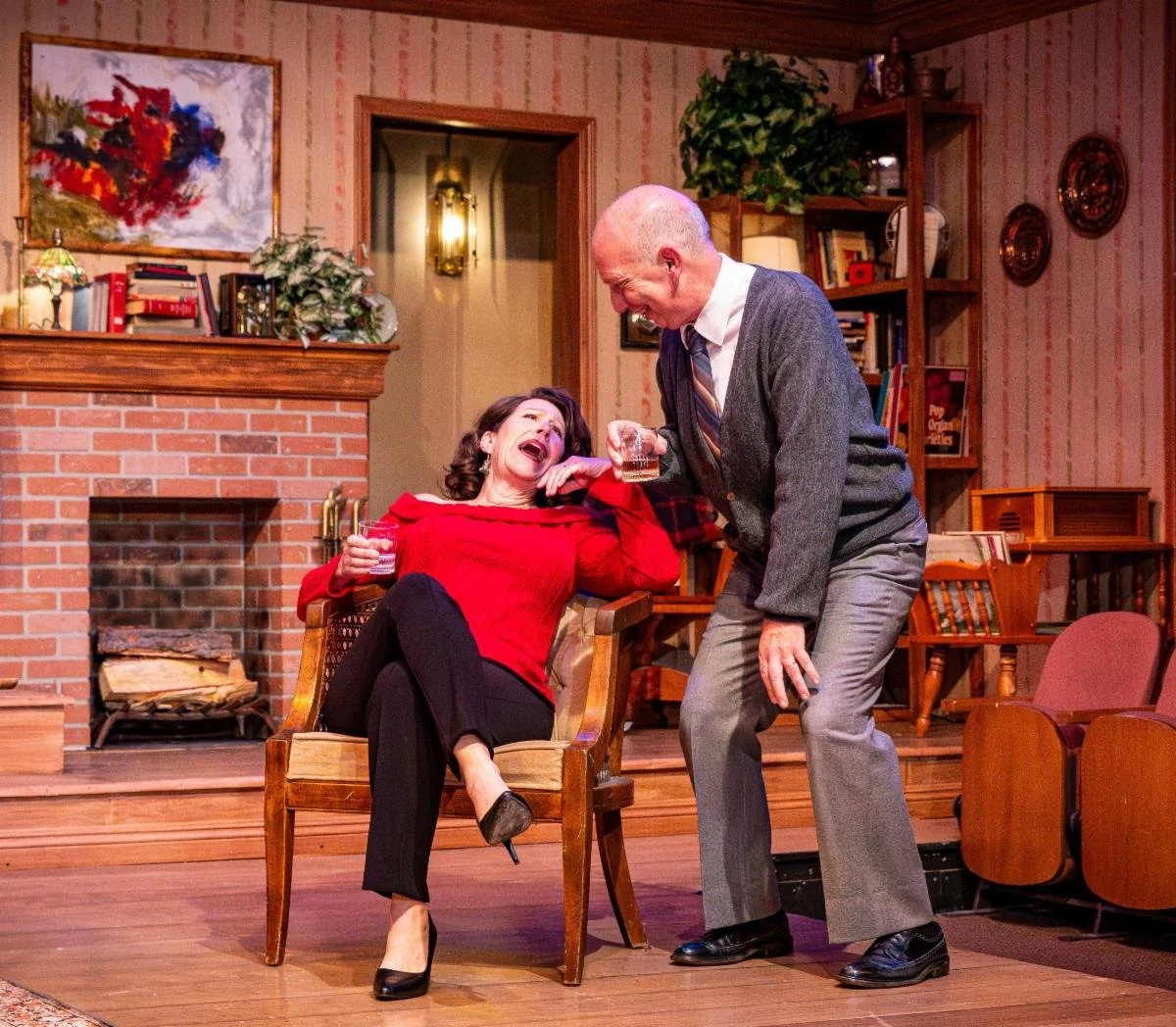 A woman in a red top and black pants is laughing while sitting in a wooden chair, holding a glass of drink. A man in gray pants, a white shirt, tie, and gray sweater is standing next to her, laughing and holding a glass. They are in a cozy living roo