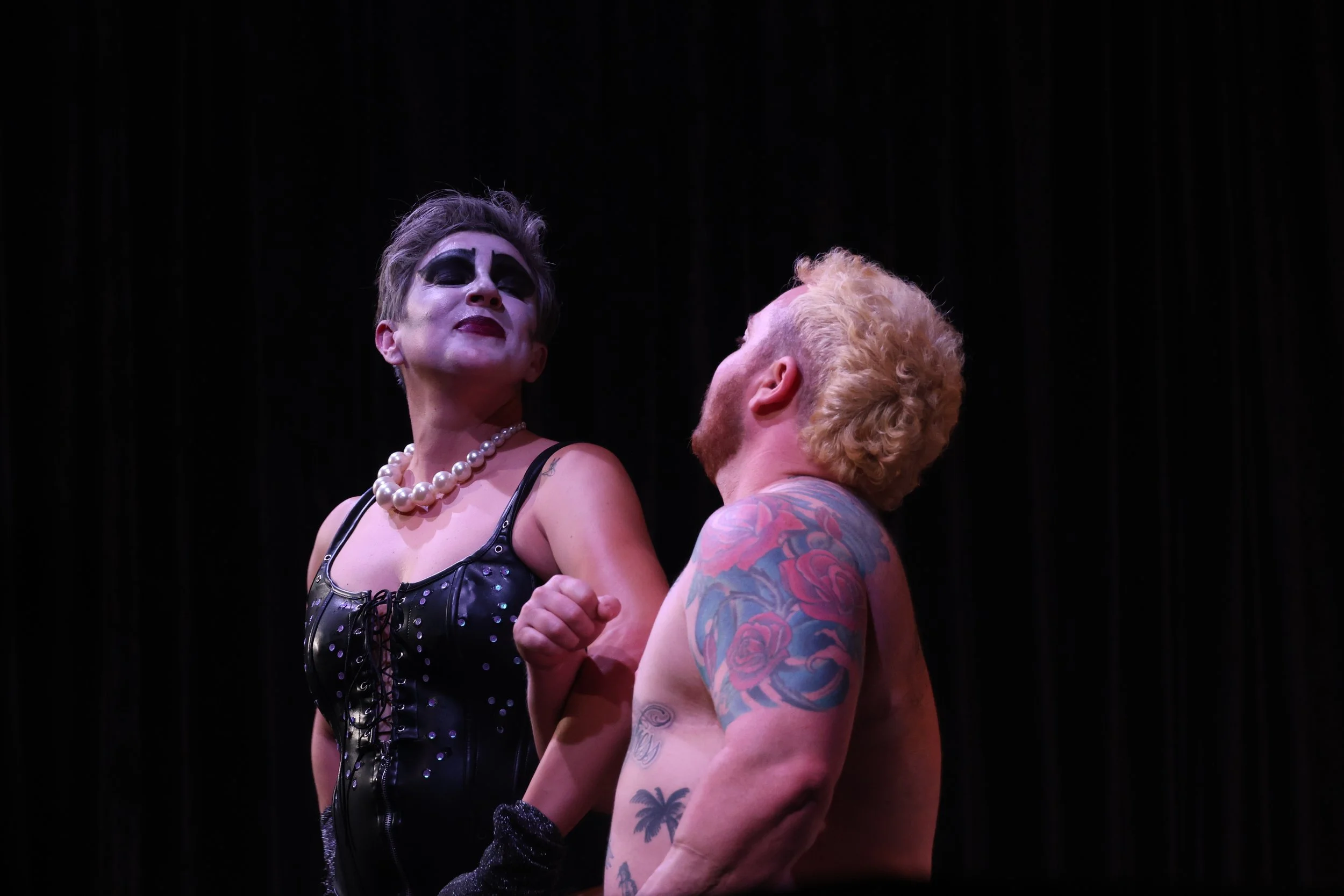 A woman dressed in black leather with pearl necklace and makeup stands closely facing a shirtless man with tattoos and red curly hair on a dark stage.