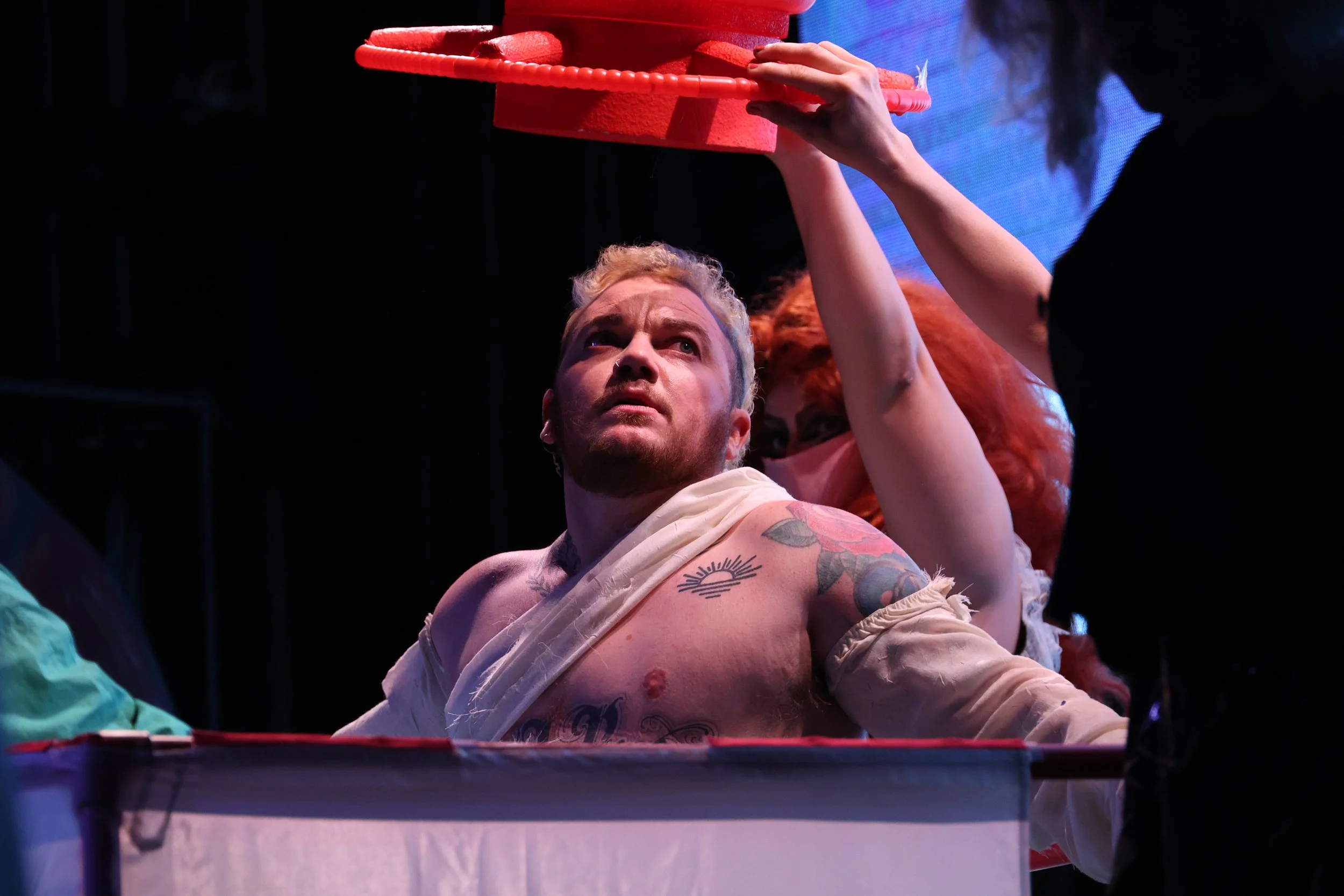 A man with tattoos leaning on a table with a serious expression, a woman with red hair holding a red, foam-like object above his head, dark background with a blue screen in the back.