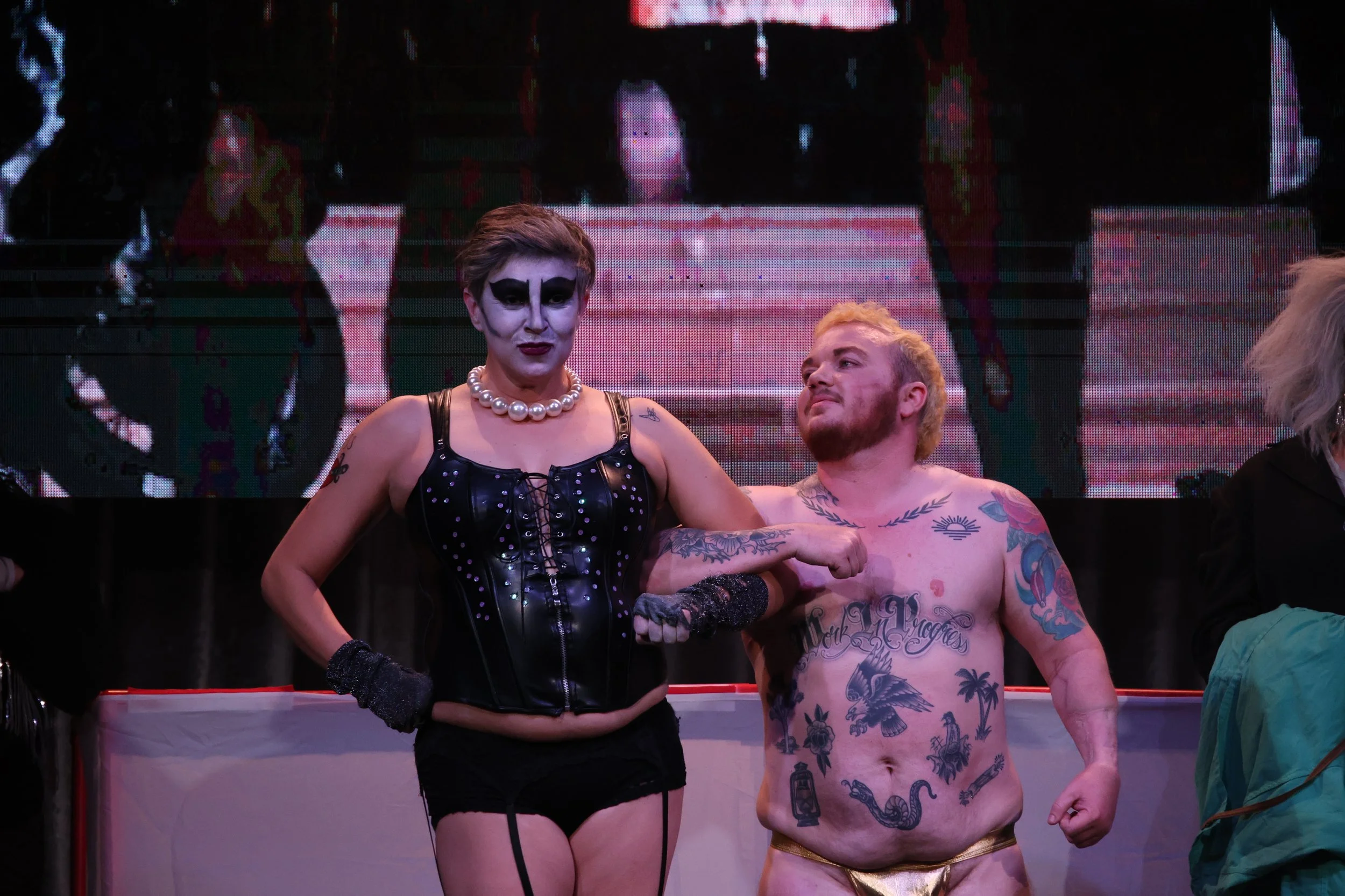 A woman with makeup and a pearl necklace standing next to a shirtless man with tattoos in a boxing ring.